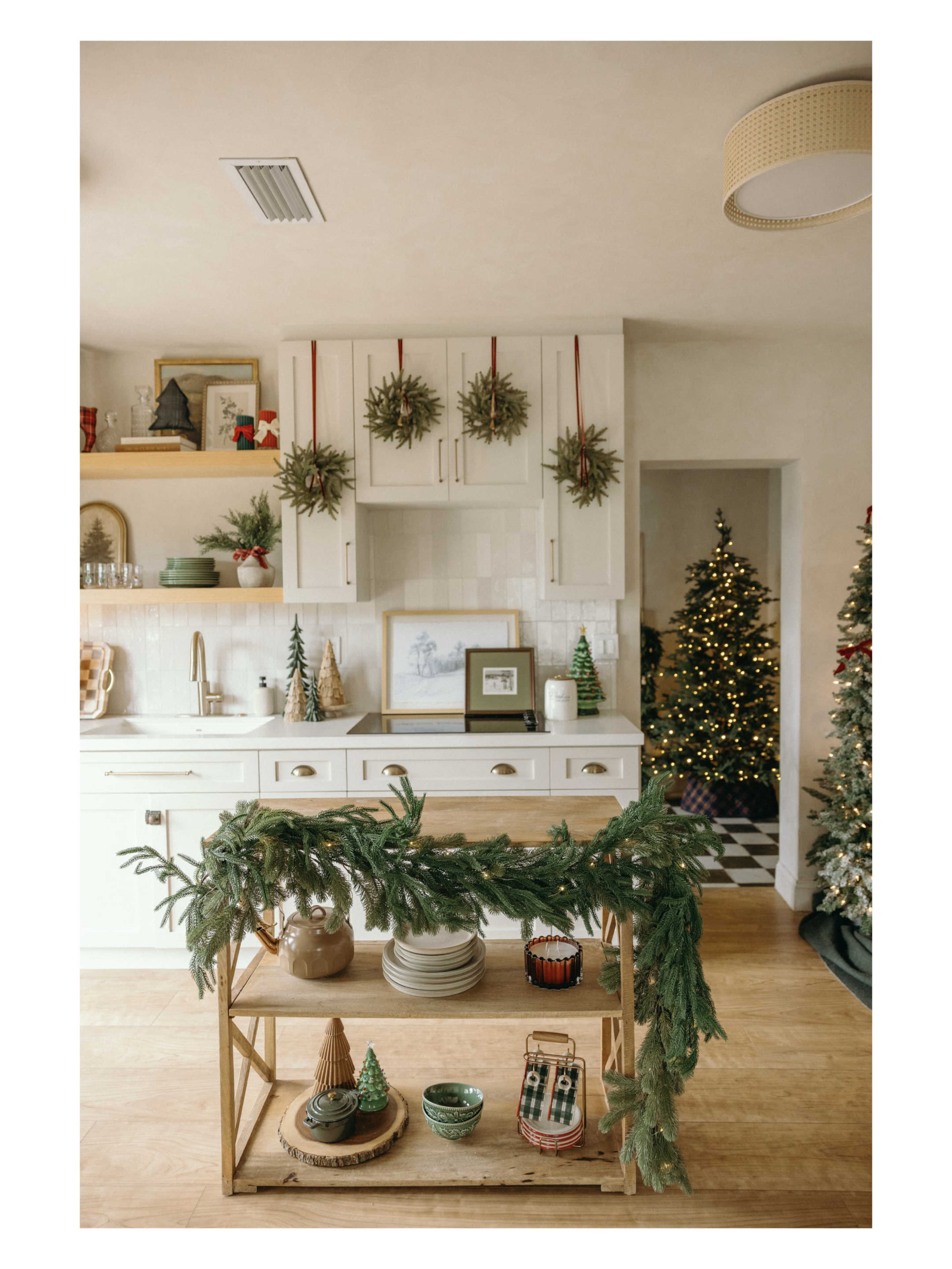 A decorated kitchen features a wooden cart with plates and festive greenery, while a Christmas tree adorned with lights stands in the background.