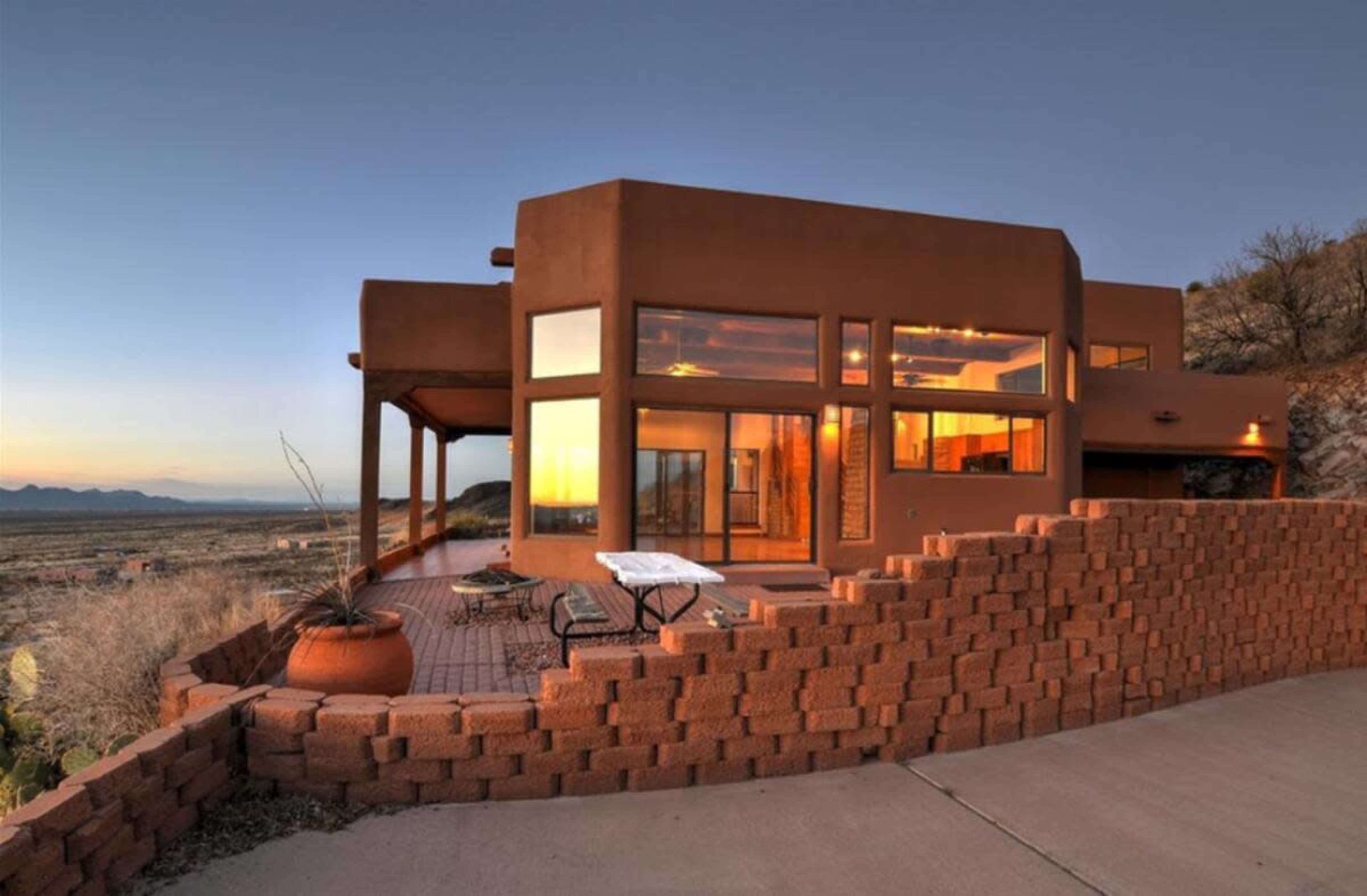A modern, rectangular adobe house with large windows overlooks a vast desert landscape at sunset.