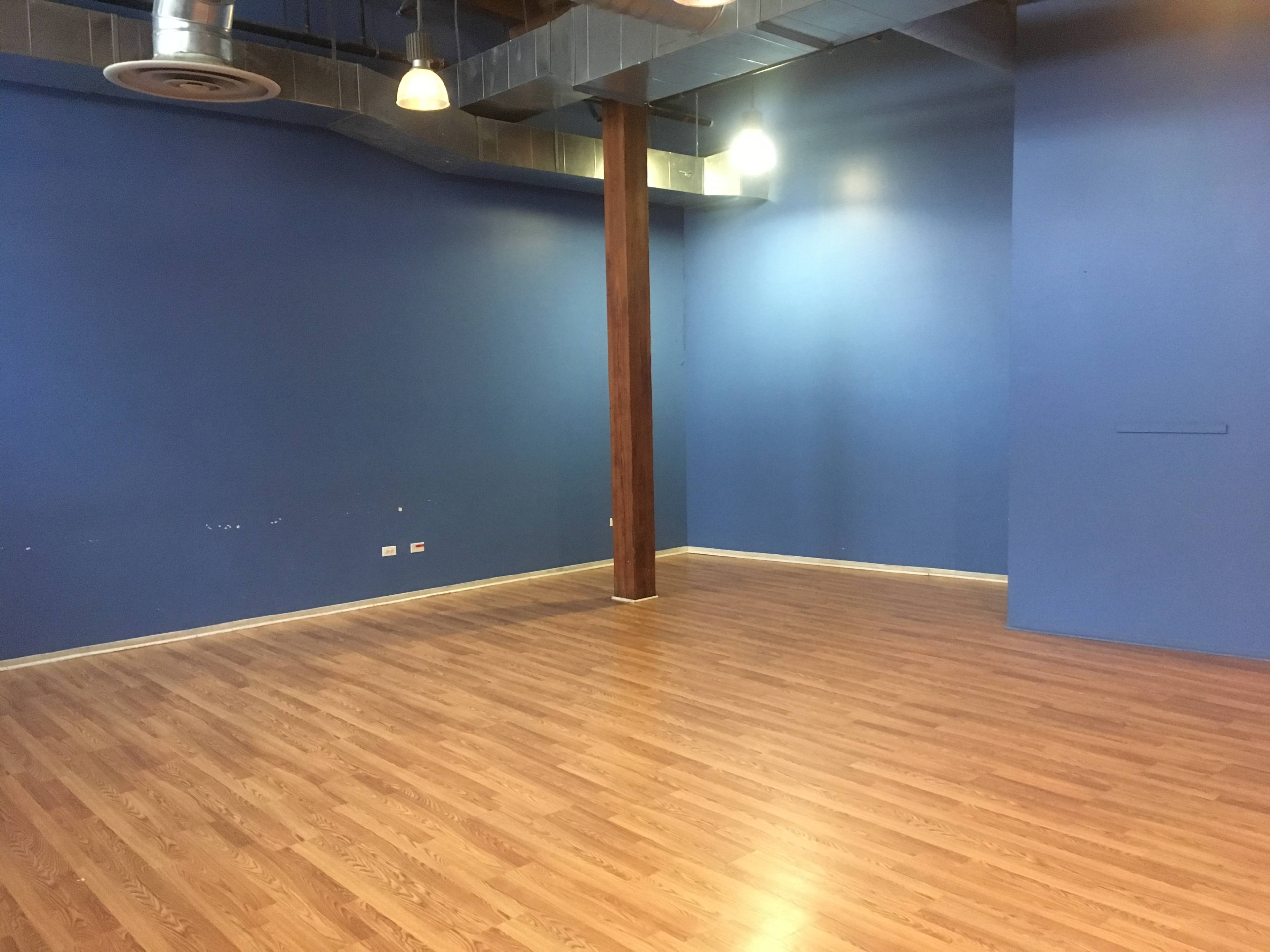 A spacious room with blue walls, wooden flooring, and exposed ductwork, featuring a central wooden column and two overhead lights.