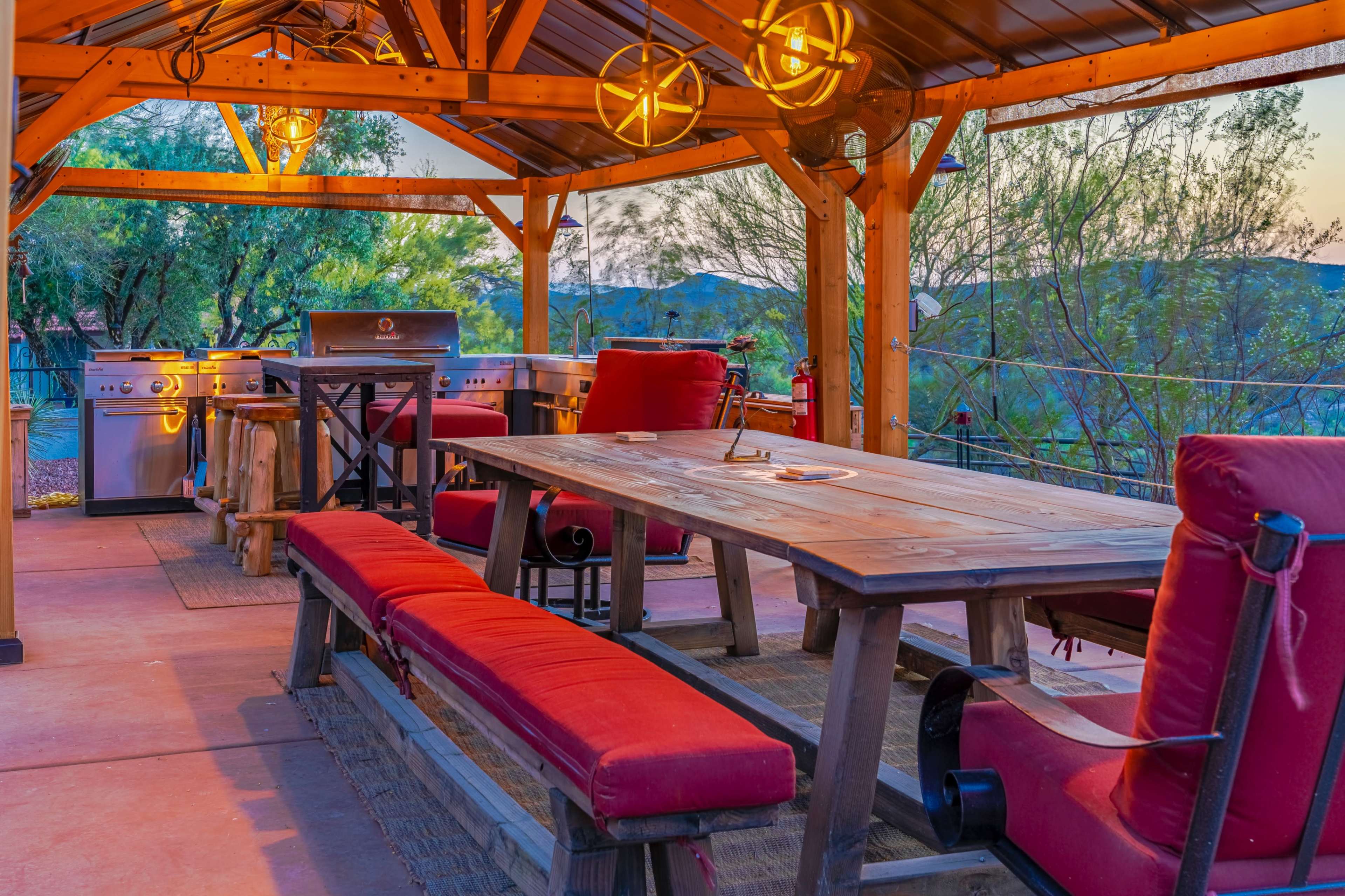 The image shows a rustic outdoor dining area with a wooden table, red cushioned seating, and a grill under a decorative gazebo illuminated by hanging lights.