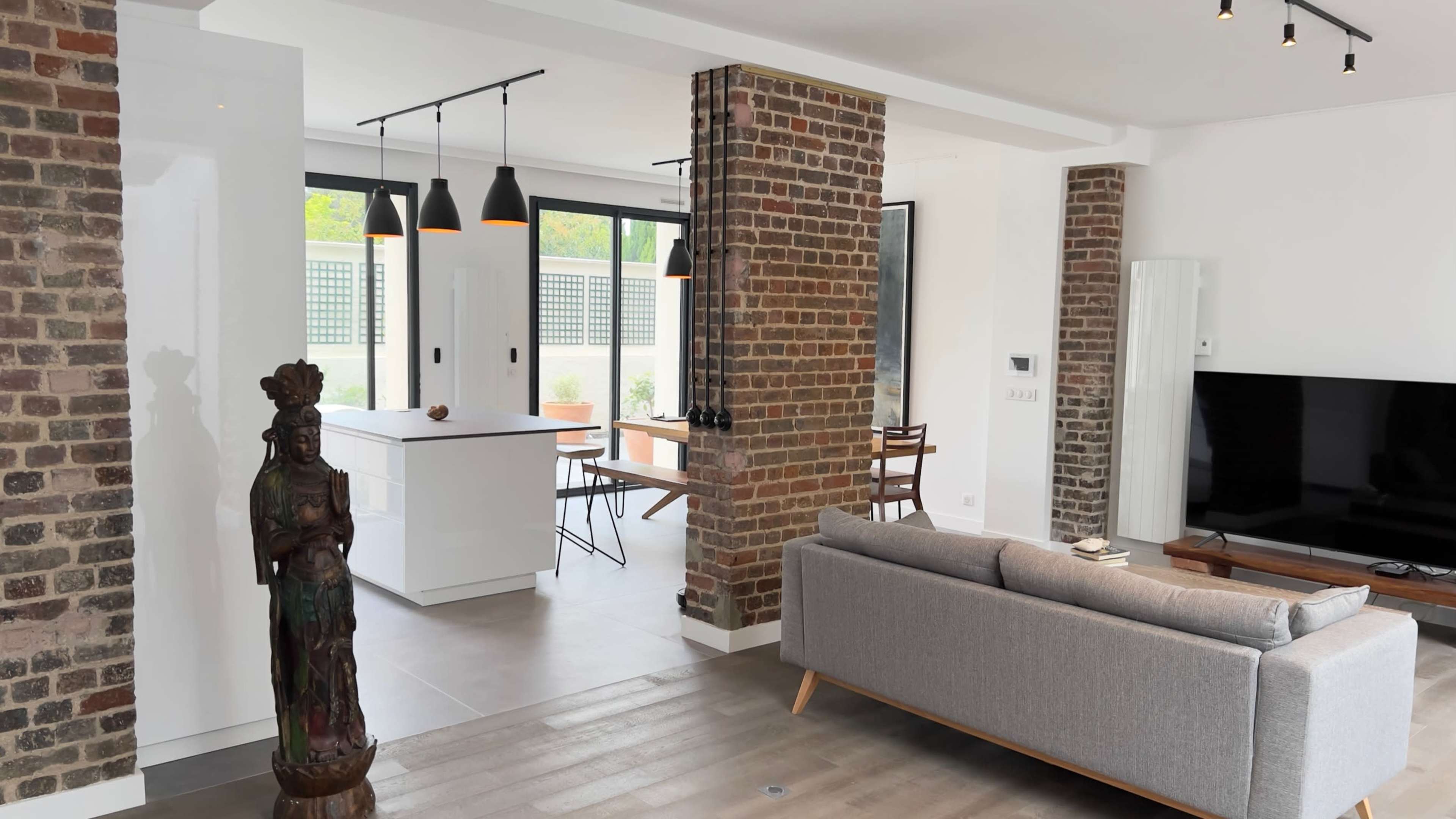 A modern living space with a gray sofa, a decorative statue, exposed brick columns, and an open kitchen area with pendant lighting.