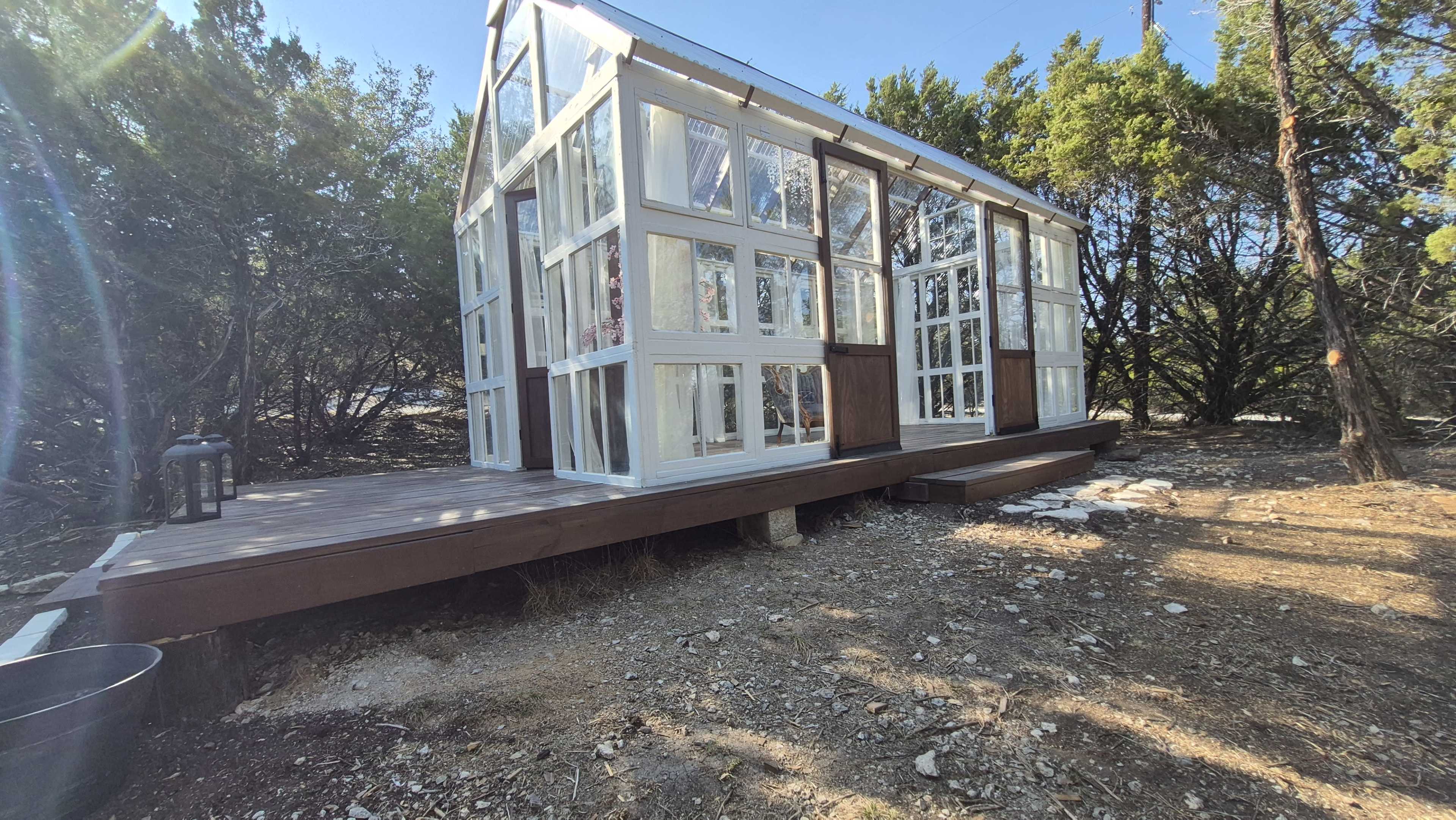 The image shows a glass greenhouse with wooden accents situated on a rocky landscape surrounded by trees.