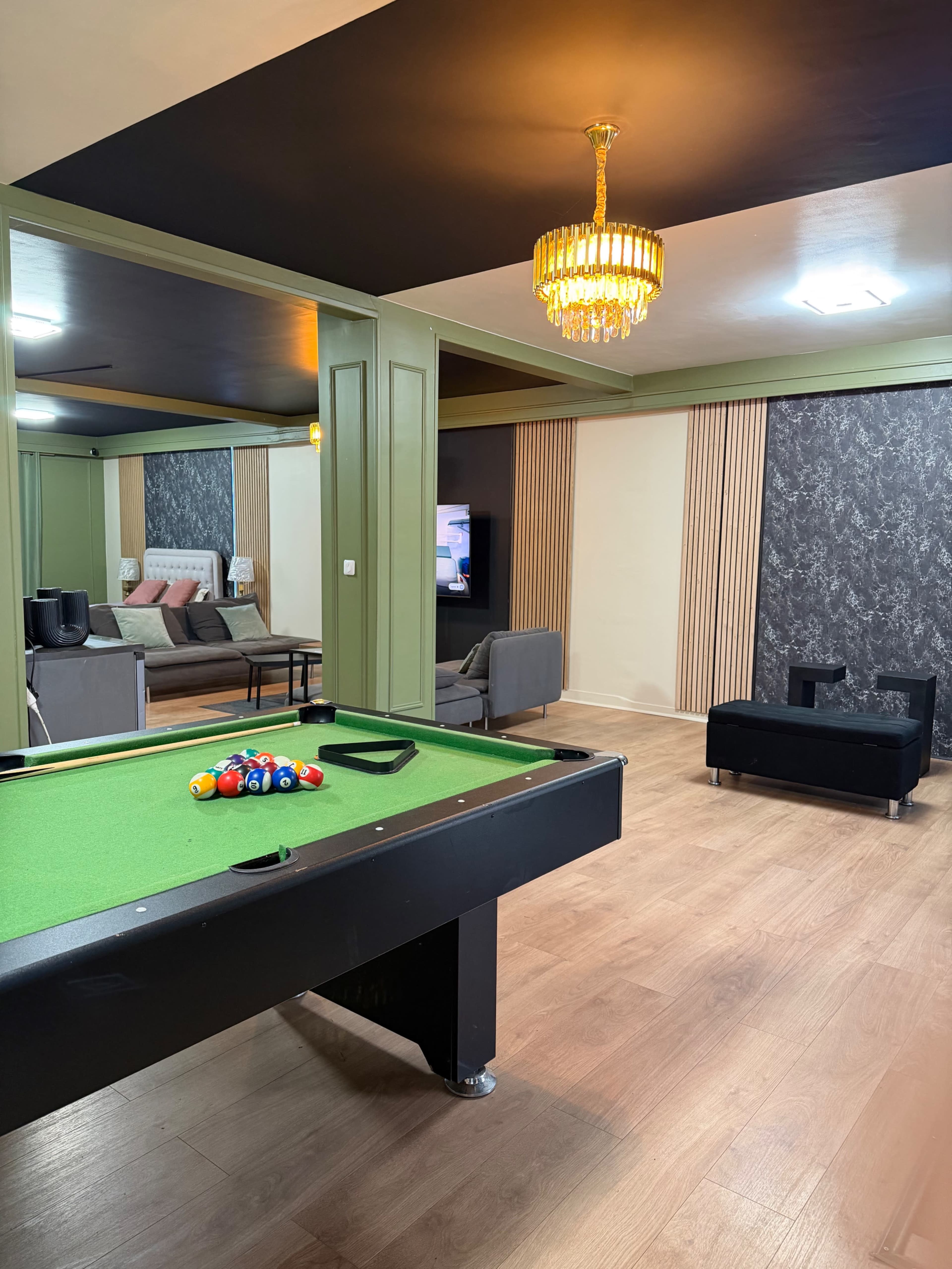 The image depicts a modern game room featuring a pool table, a seating area with a sofa, and a television against a textured accent wall.