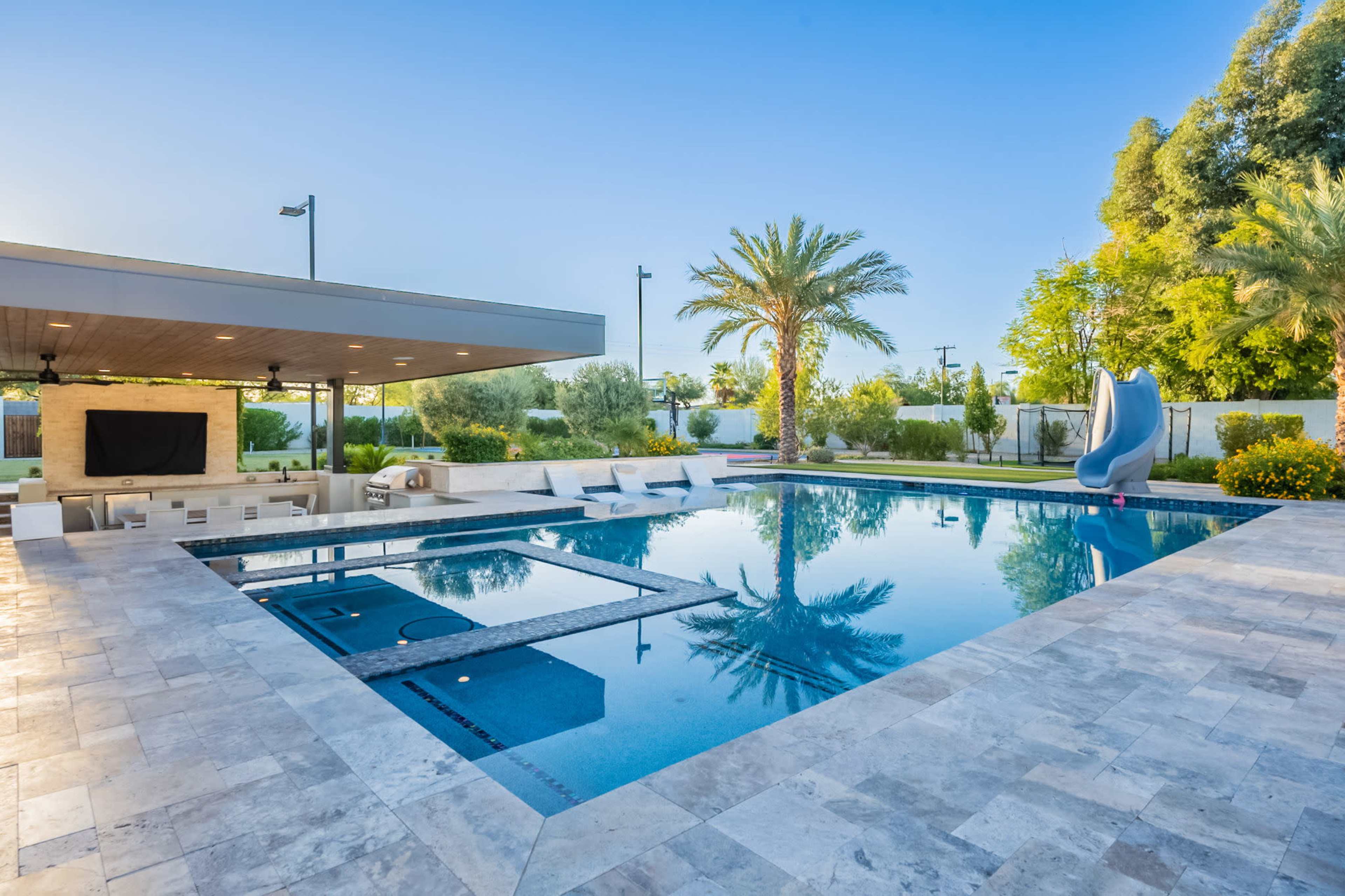 The image shows a modern outdoor pool area featuring a large swimming pool, a slide, lounge chairs, and palm trees under a clear blue sky.