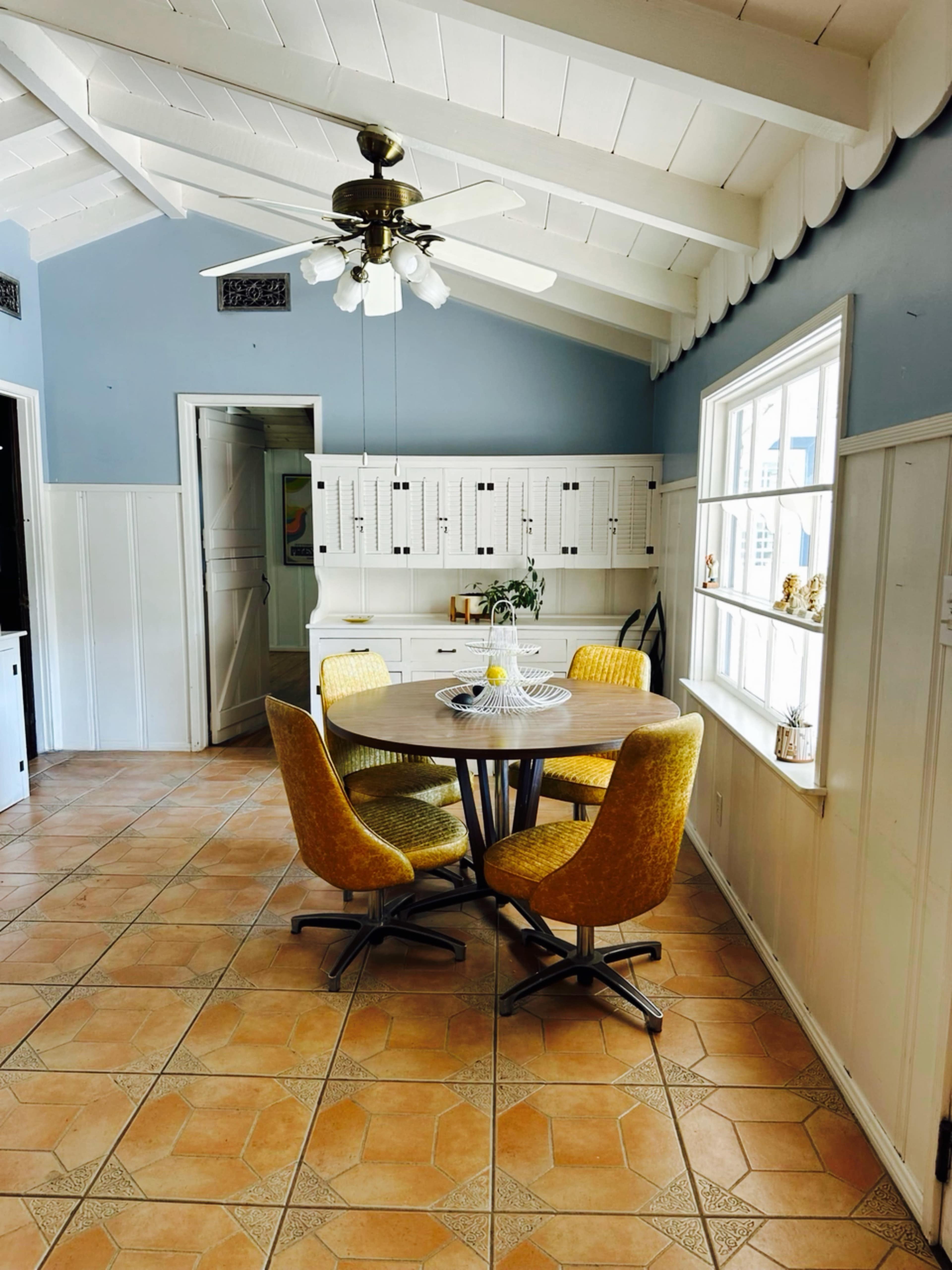 A round wooden table surrounded by four yellow chairs sits in a bright kitchen with white walls and a ceiling fan.
