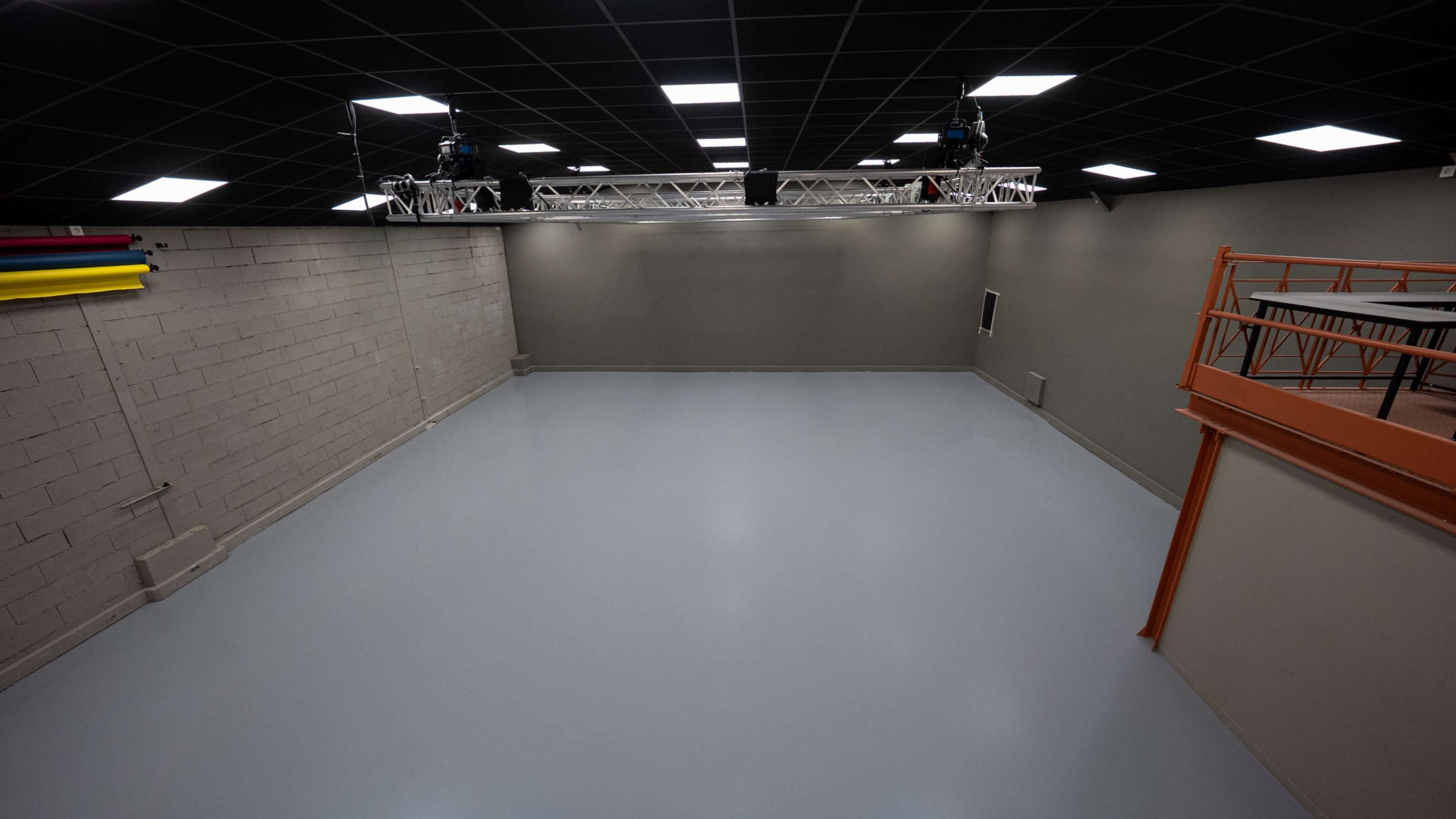 The image shows an empty, spacious studio with gray walls and a smooth light-colored floor, illuminated by overhead lights and equipped with overhead rigging.