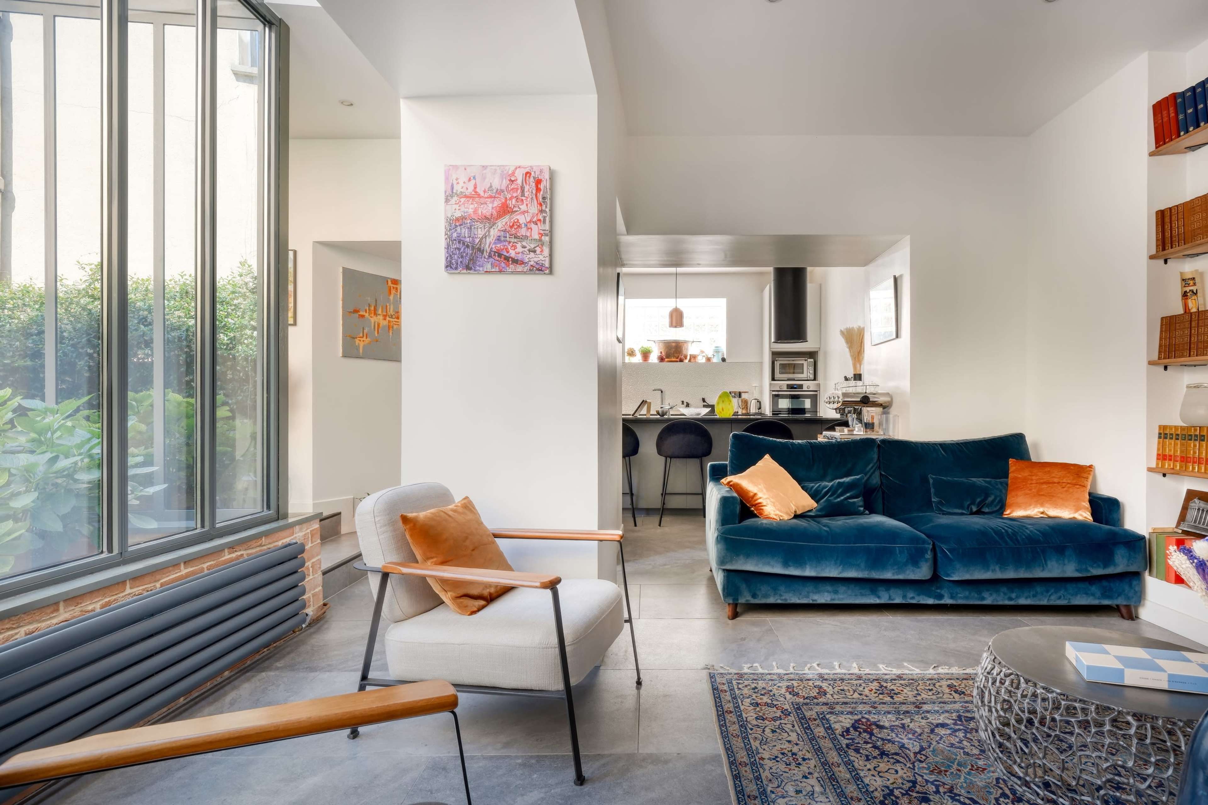 The image shows a modern living space featuring a blue velvet sofa, a light-colored armchair, and a visible kitchen area in the background.