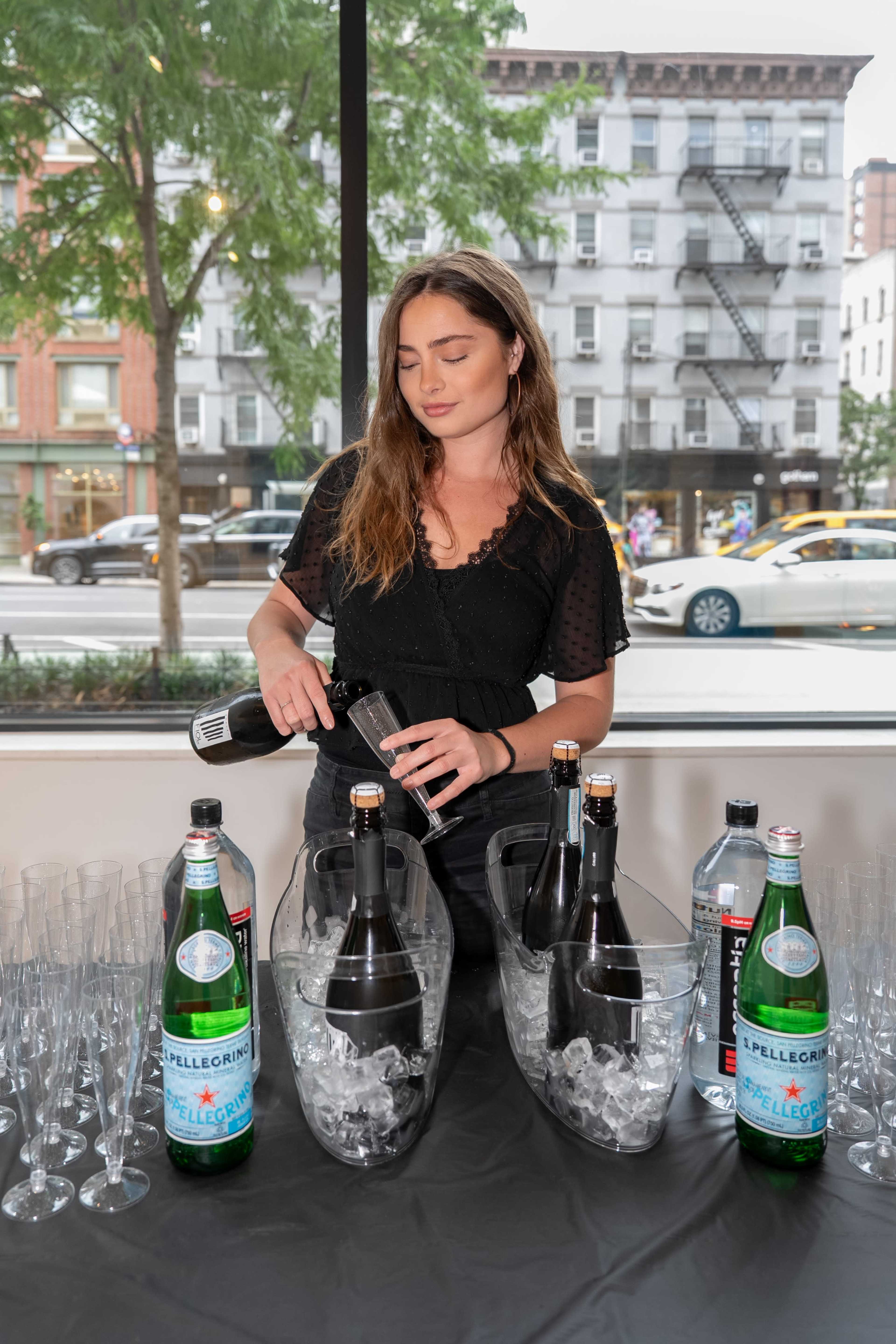 A woman is pouring a beverage from a bottle into a glass at a table with bottles of sparkling water and champagne, surrounded by ice and champagne flutes.