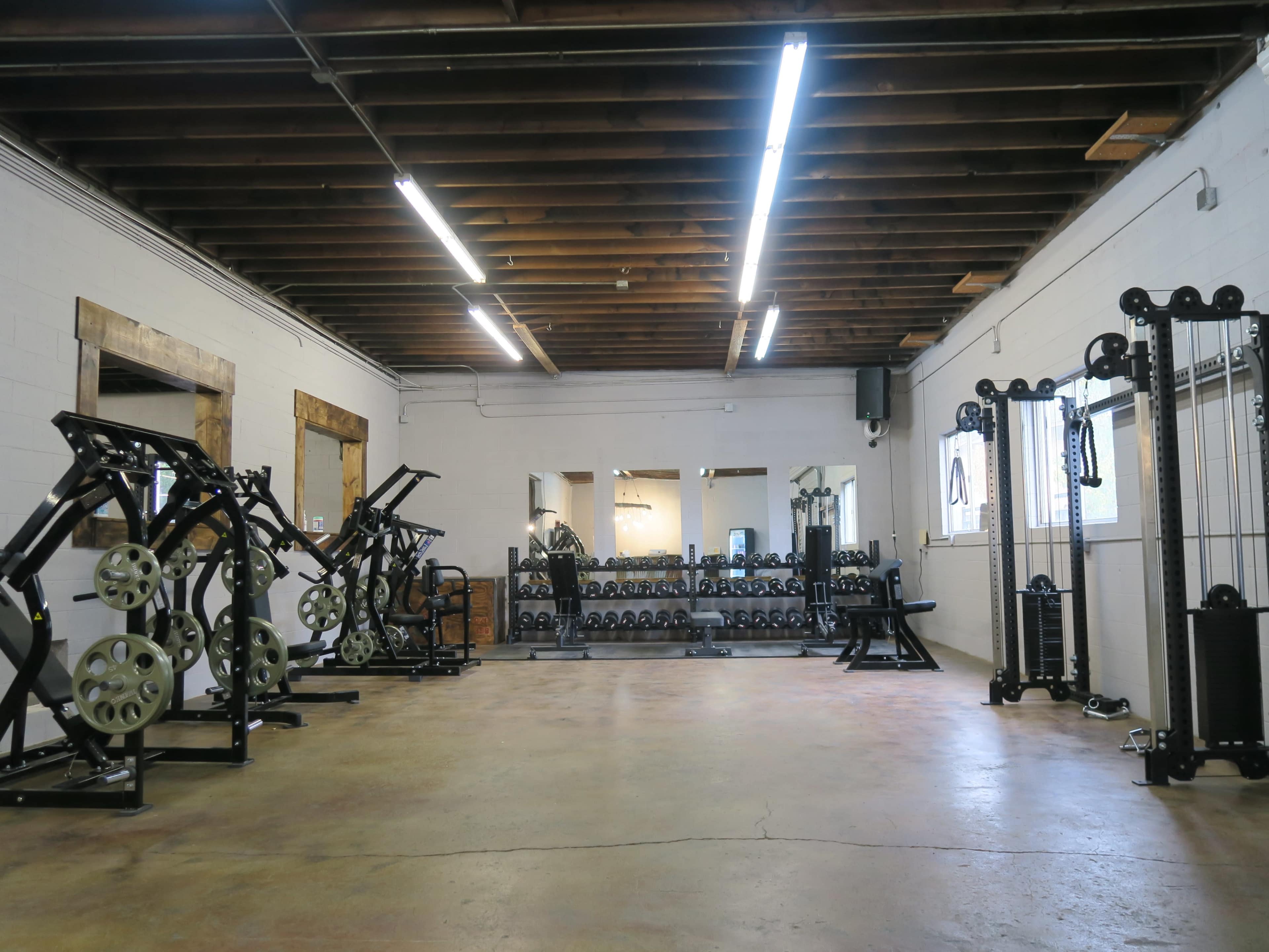 A spacious gym with various weightlifting equipment, including dumbbells, cable machines, and squat racks, under a wooden ceiling.