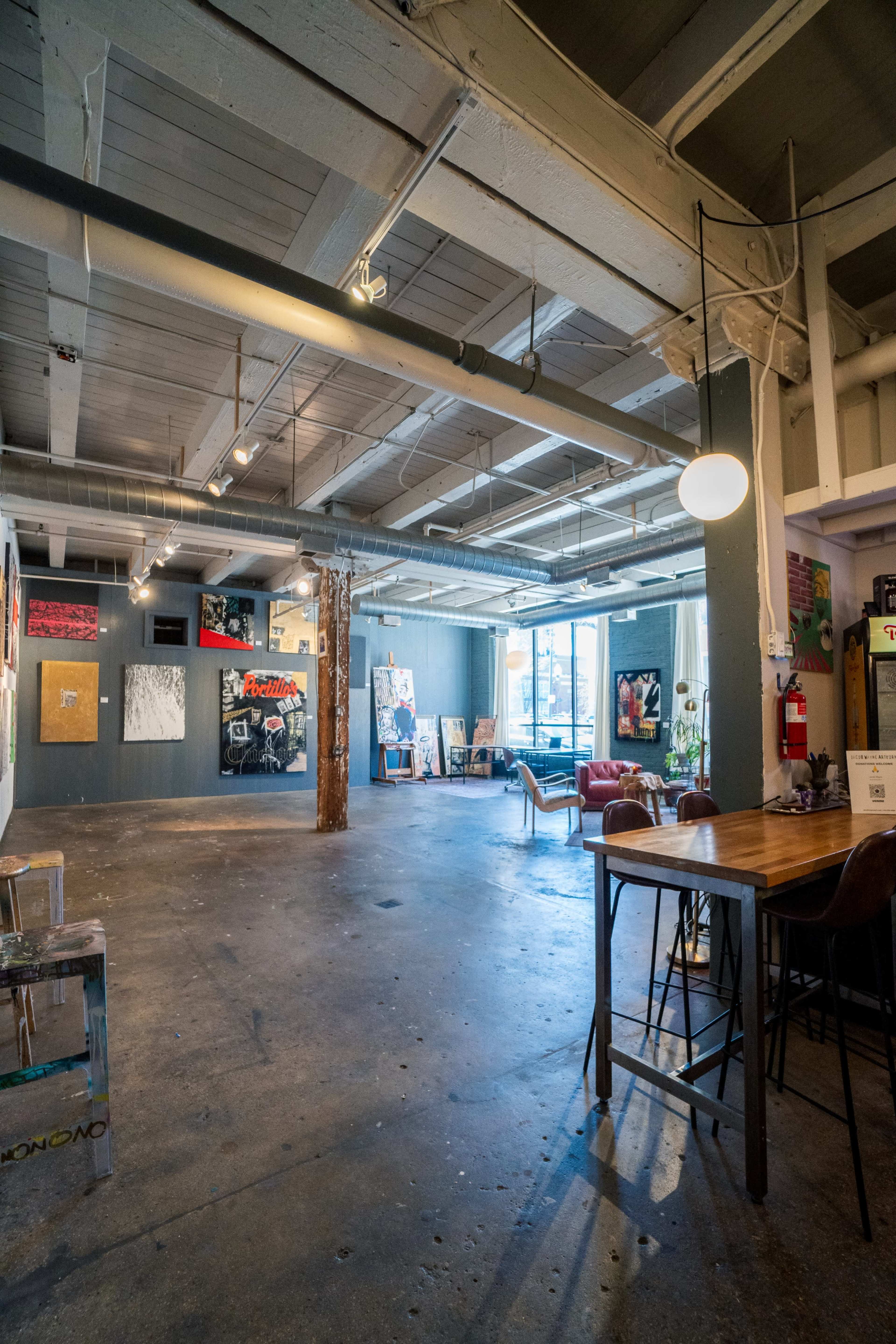 A spacious, industrial-style art gallery with exposed beams, wooden tables, and various artworks displayed on the walls.