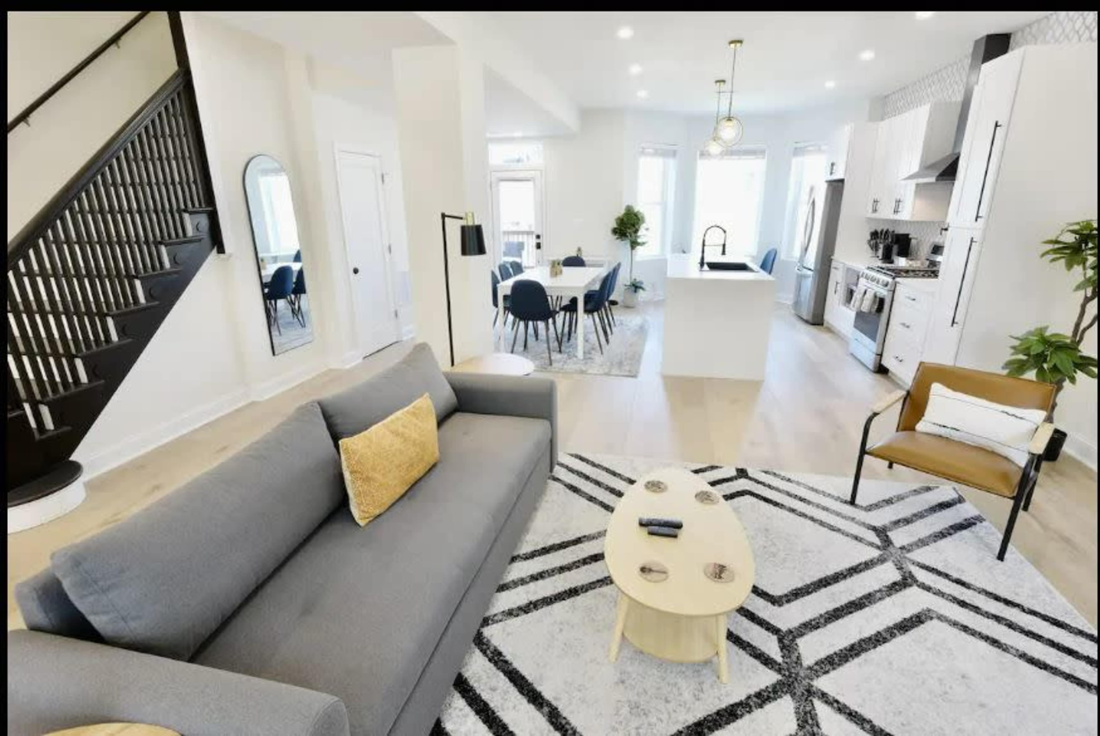 The image shows a modern living room featuring a gray sofa, a round coffee table, and a staircase, leading to a dining area with a kitchen in the background.