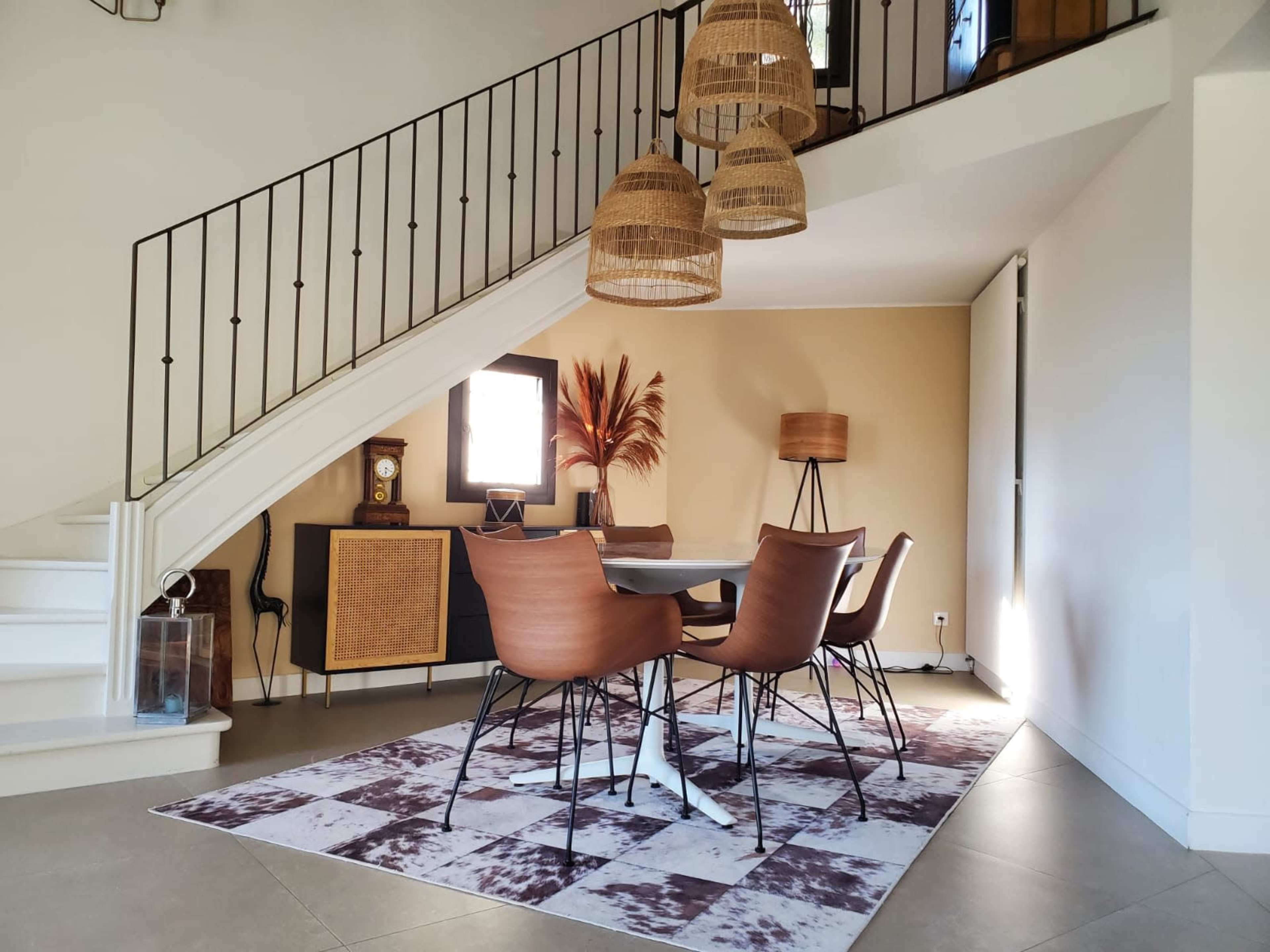 The image shows a modern dining area with a round table surrounded by chairs, situated beneath a staircase with woven pendant lights and decorative elements on the walls.