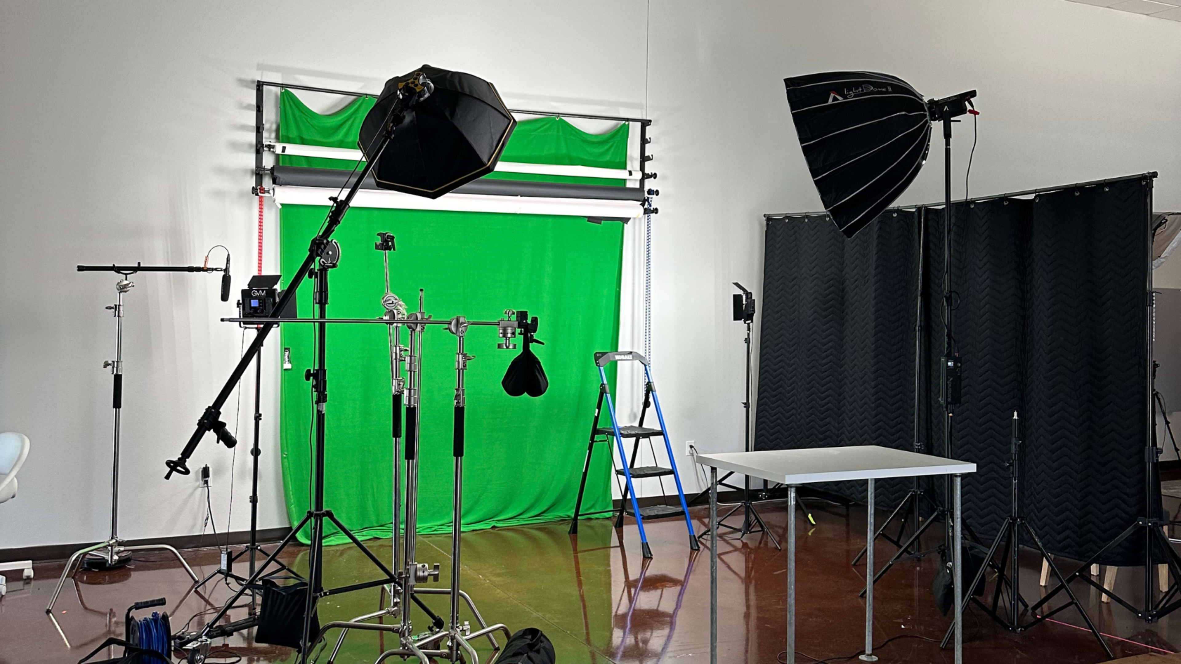 A photography studio setup with a green backdrop, lighting equipment, a ladder, and various stands.