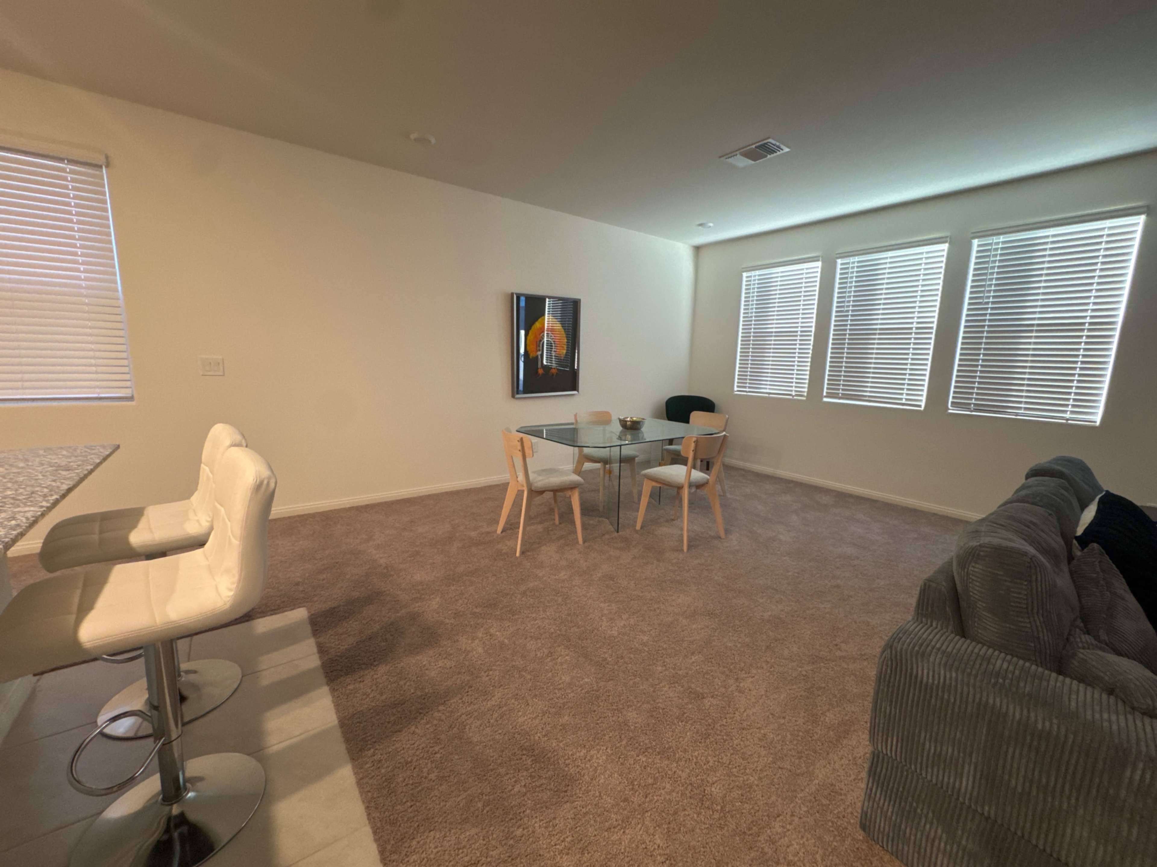 The image shows a spacious living area with a carpeted floor, a glass dining table, a gray couch, and large windows with blinds.