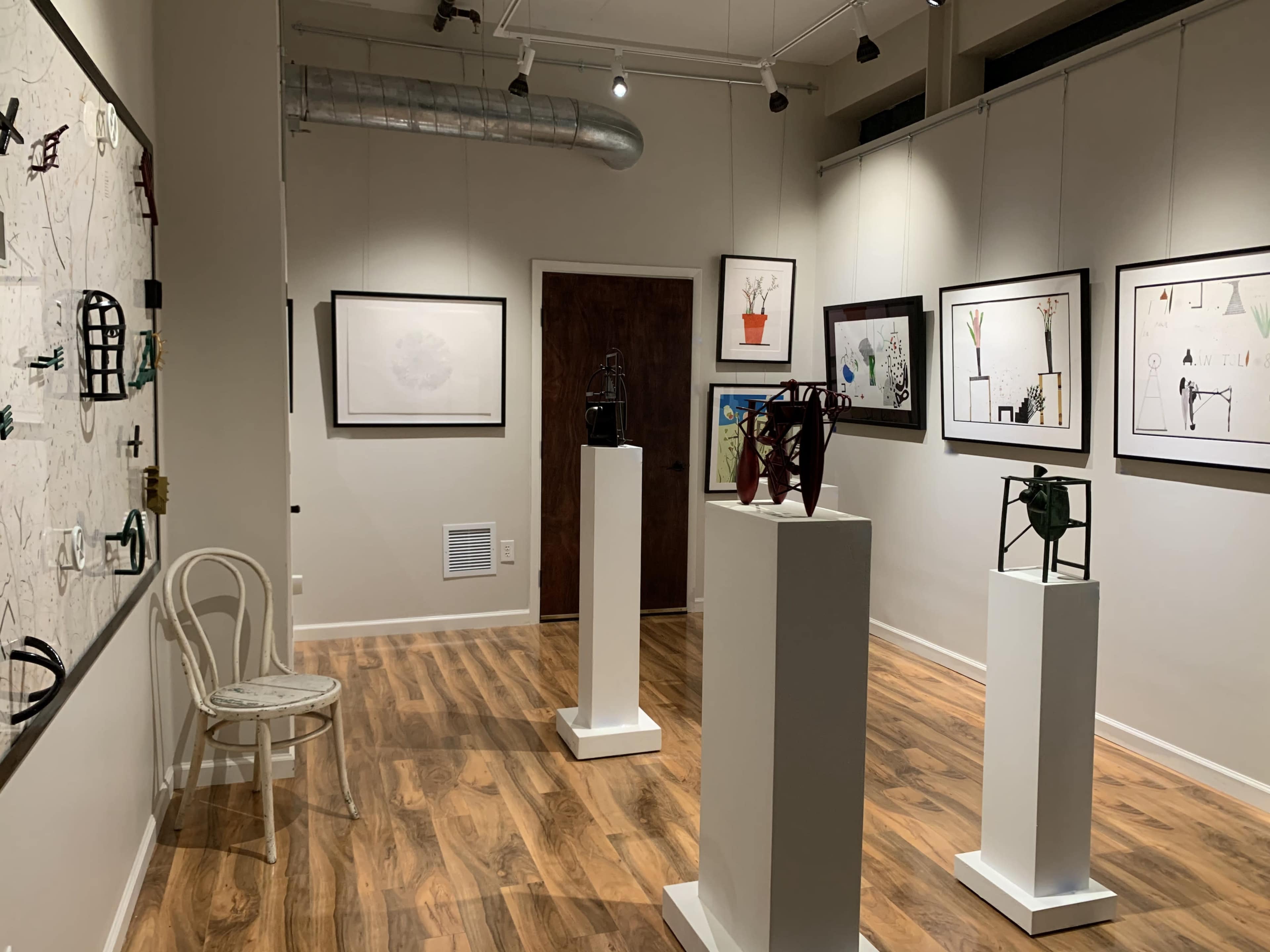 The image shows an art gallery with several framed artworks displayed on the walls and white pedestals featuring sculptures in a room with wooden flooring.