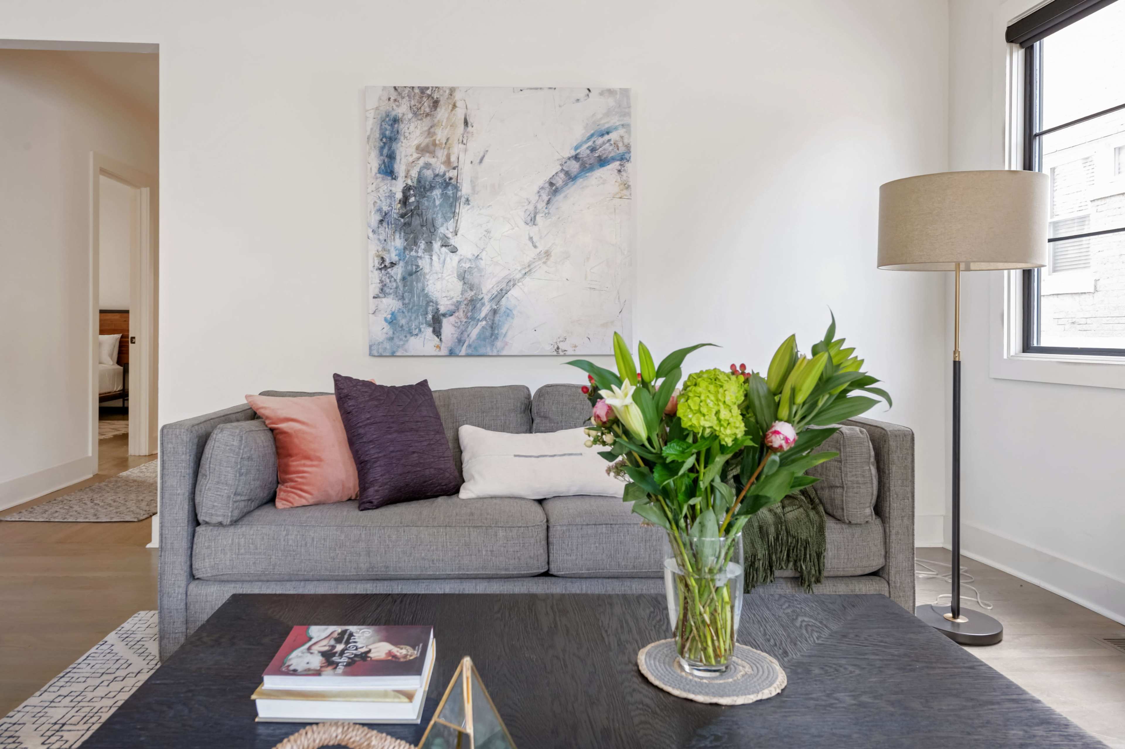 A gray sofa with decorative pillows sits in a modern living room, accompanied by a coffee table featuring books and a floral arrangement.