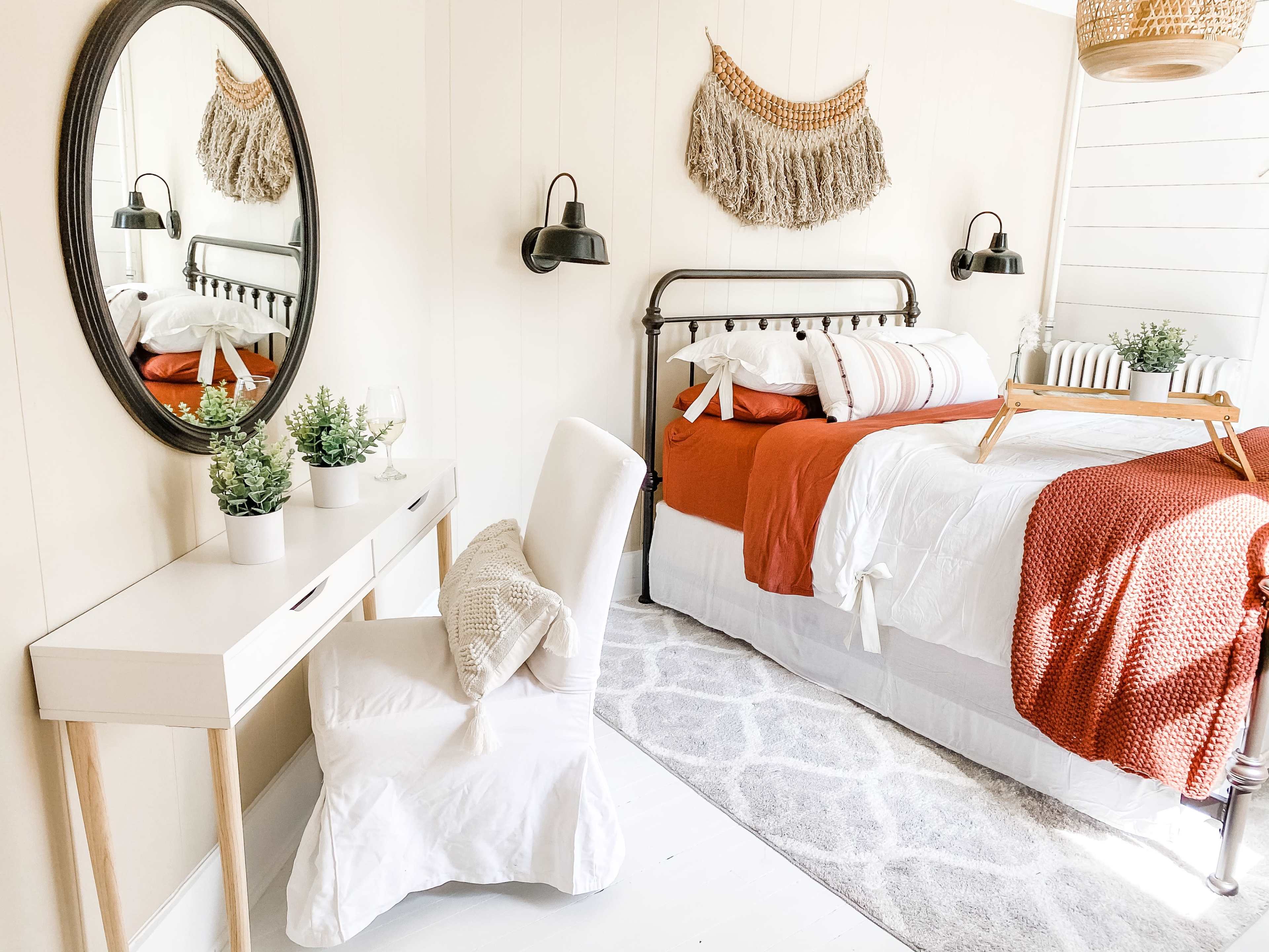 A cozy bedroom features a metal bed with red and white bedding, a round mirror above a white desk, and decorative plants on the desk and bedside.