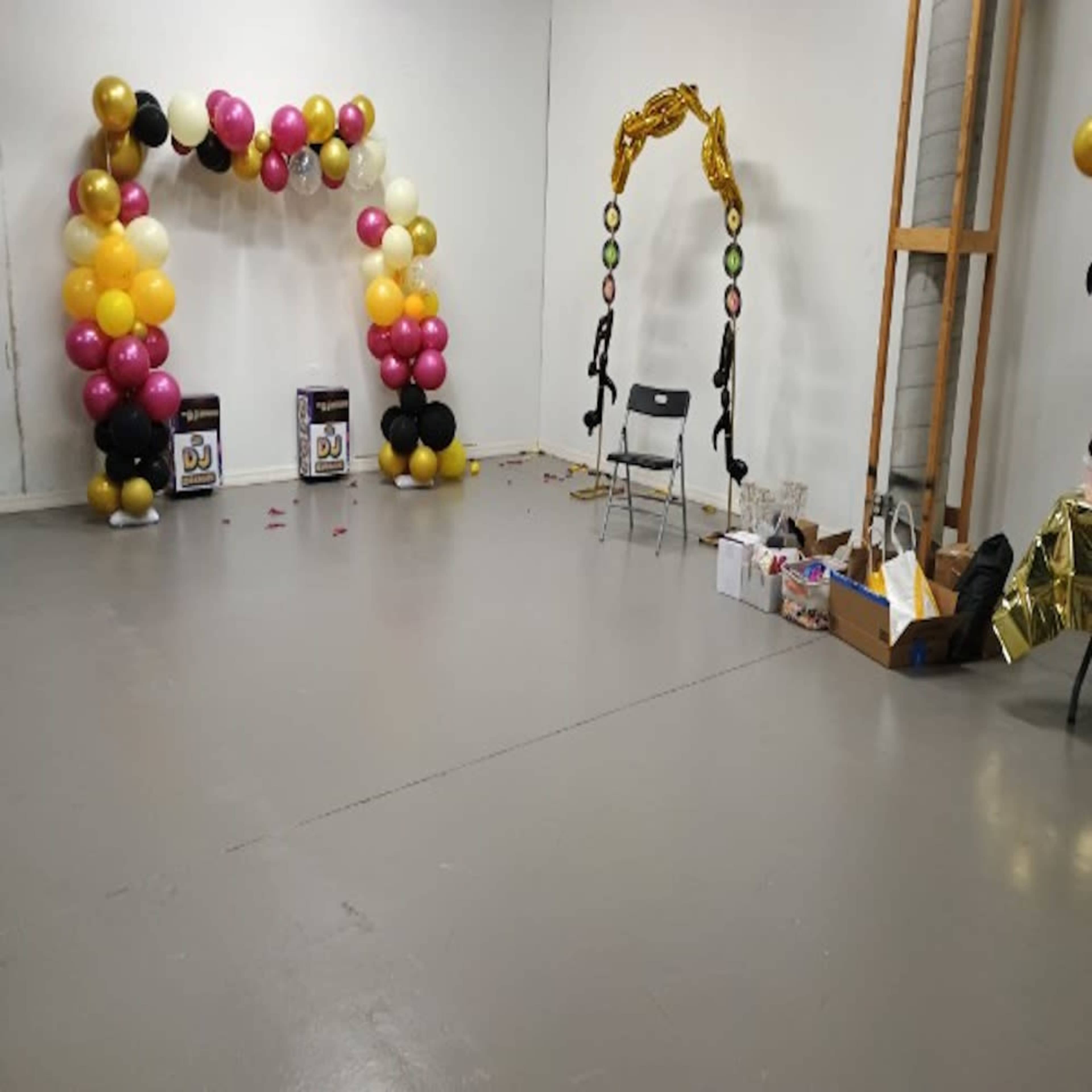 The image shows a sparse room decorated with two balloon arches in varying colors and a single chair, with scattered decorations on the floor.