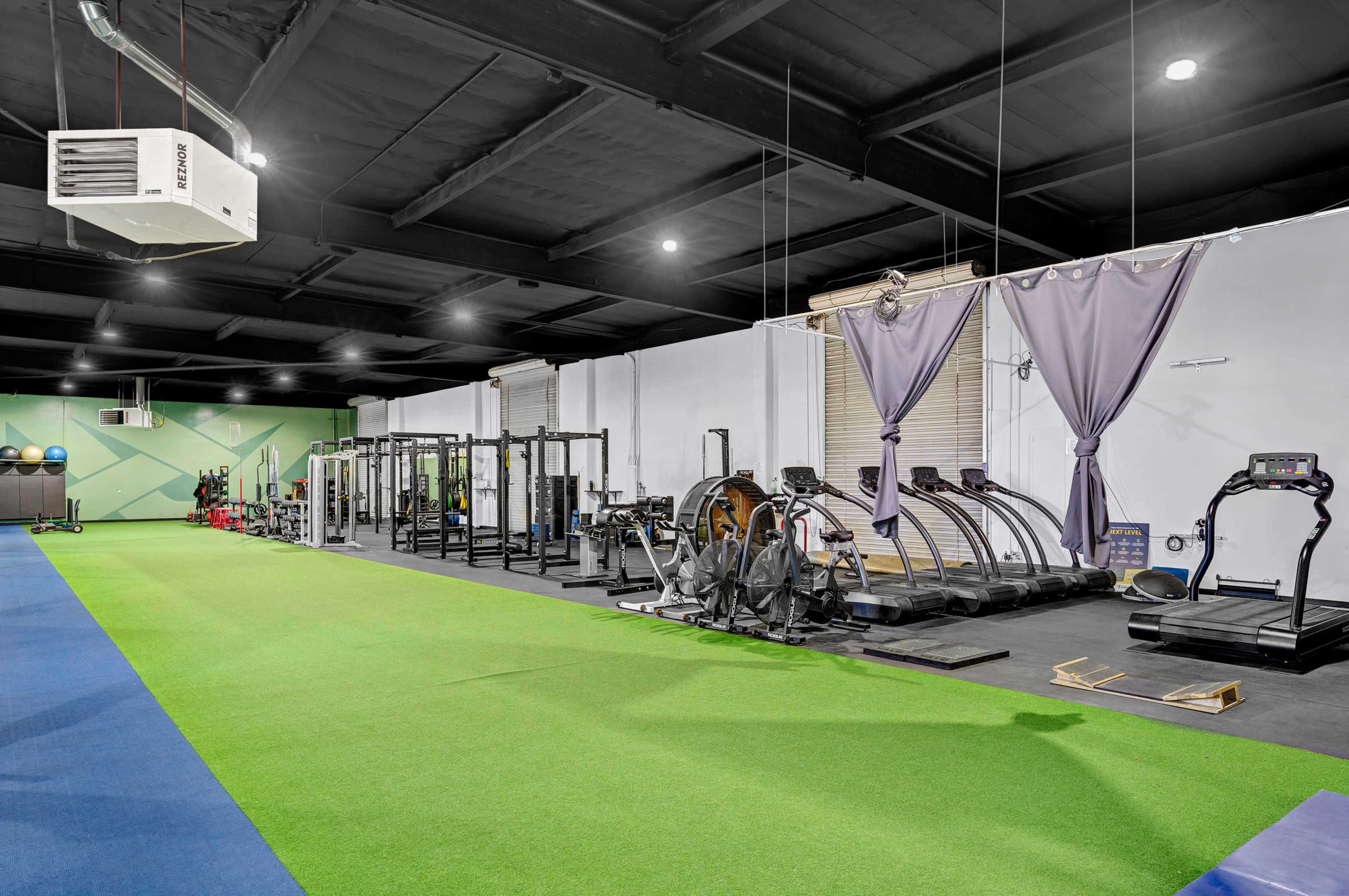 A modern gym with green turf flooring, various exercise equipment, and a section featuring treadmills and other fitness machines.