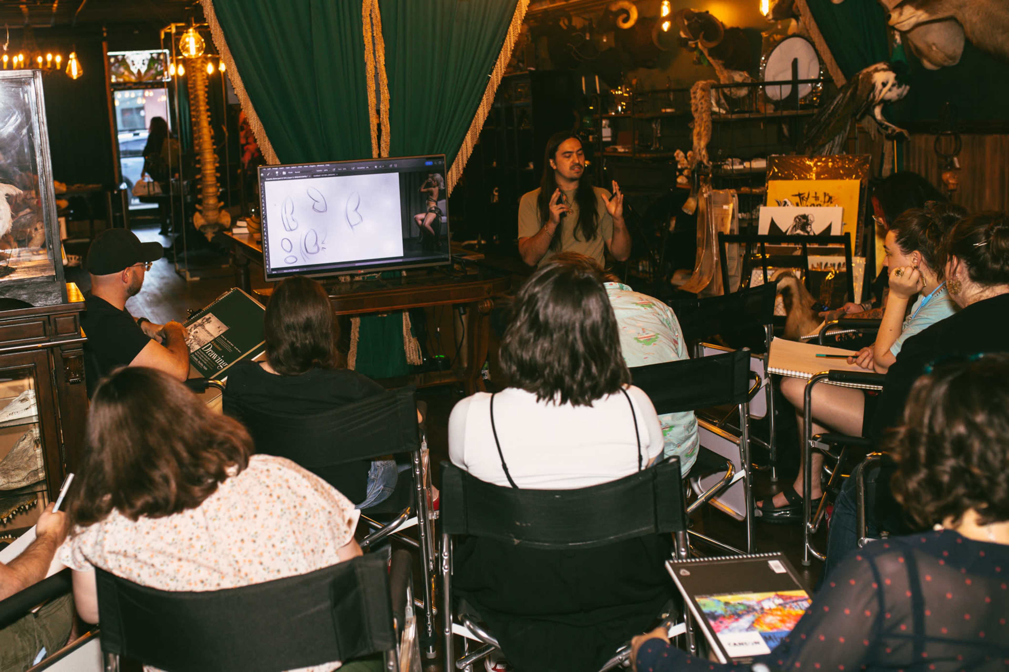 A group of people sits in chairs facing a presenter who is speaking in front of a screen displaying drawings.