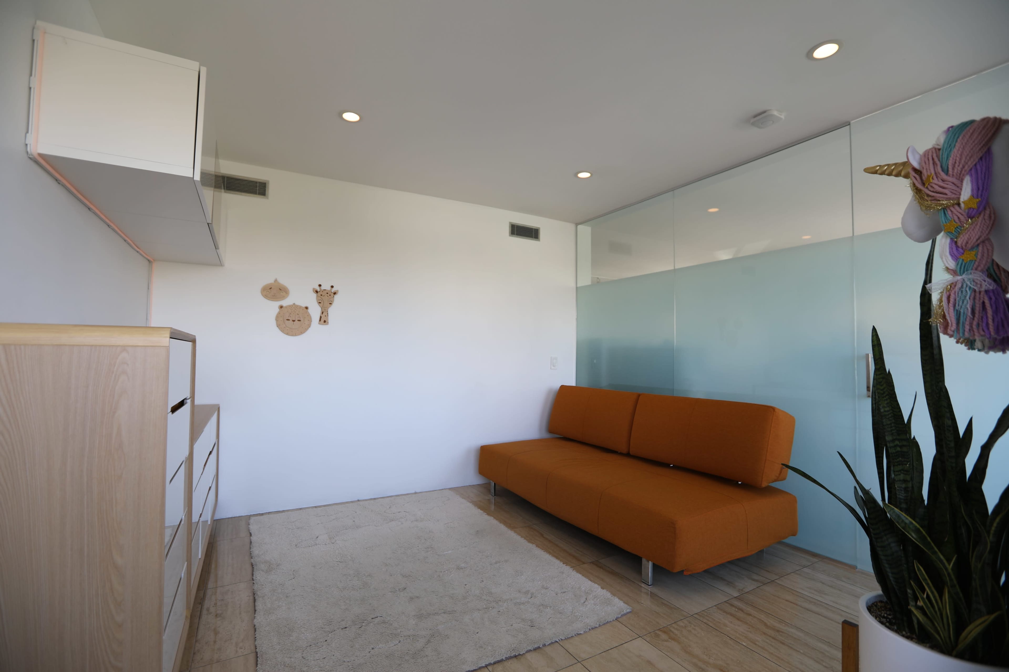 The image shows a modern, minimalist living space with an orange sofa, a rug on tiled flooring, and decorative wall hangings near a glass partition.
