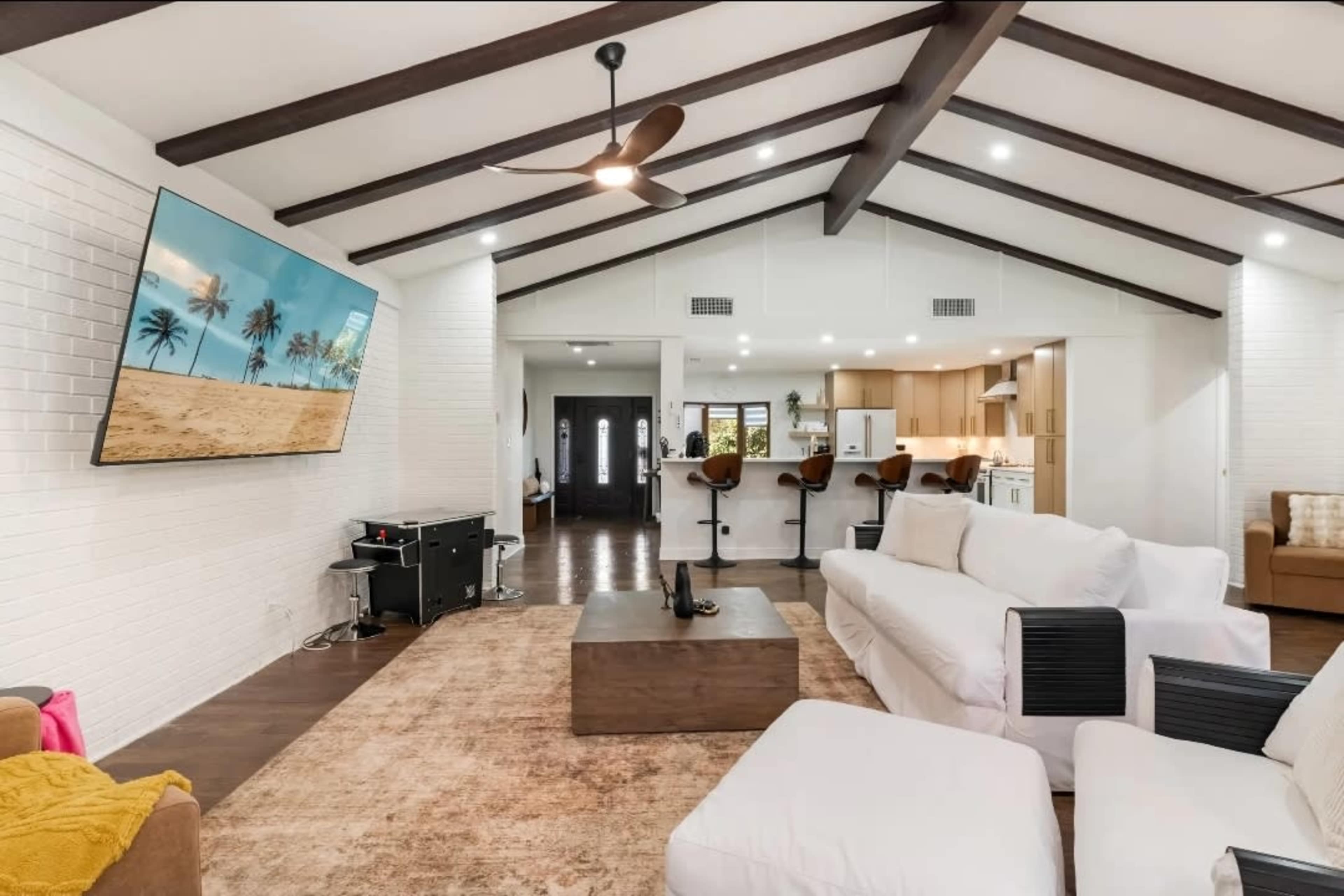 The image shows a spacious living room with a high ceiling, featuring a large TV mounted on the wall, a coffee table in the center, and seating arranged around it.
