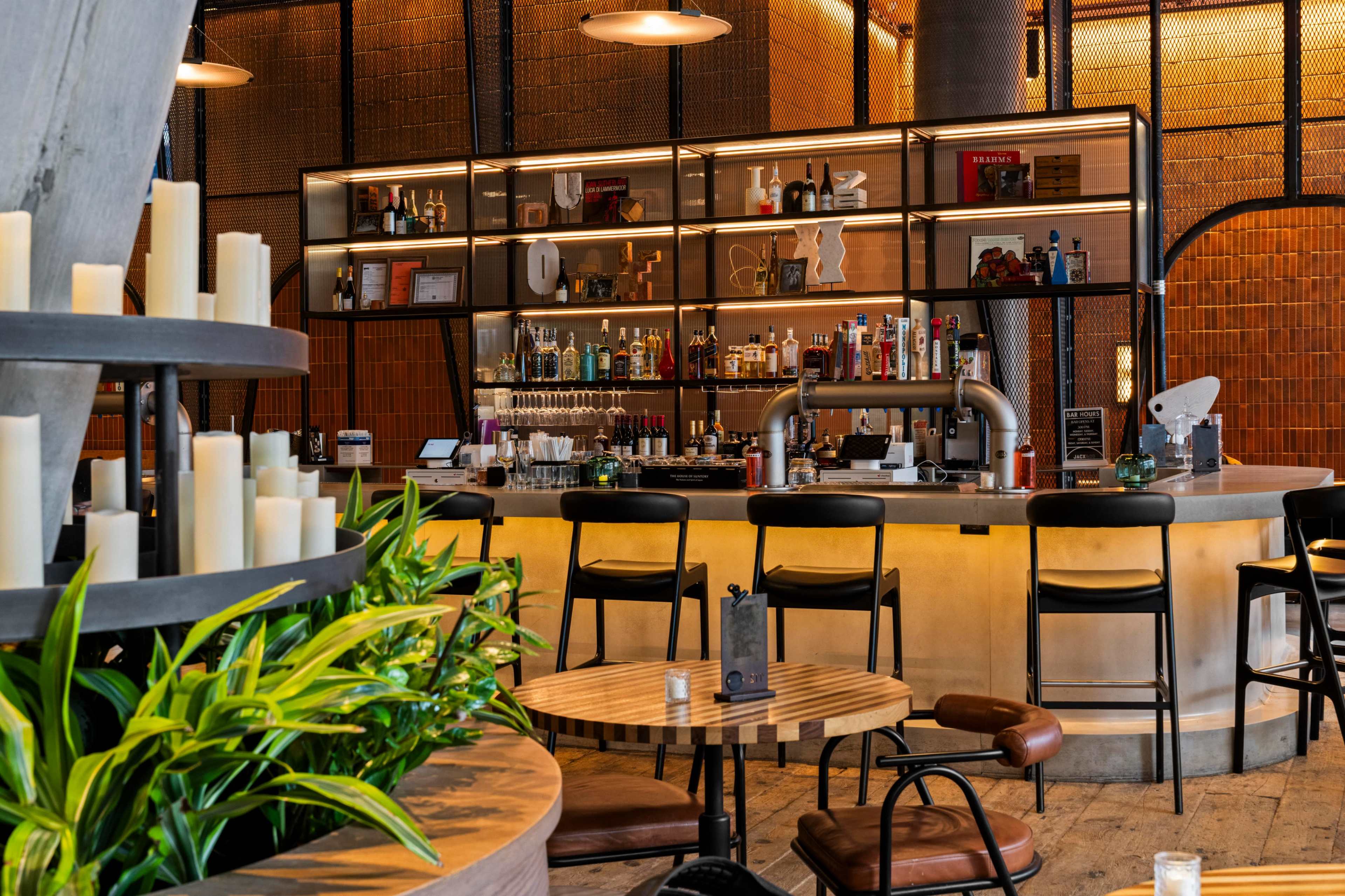 The image shows a modern bar area with a well-stocked shelf of bottles, surrounded by high stools and a few tables.