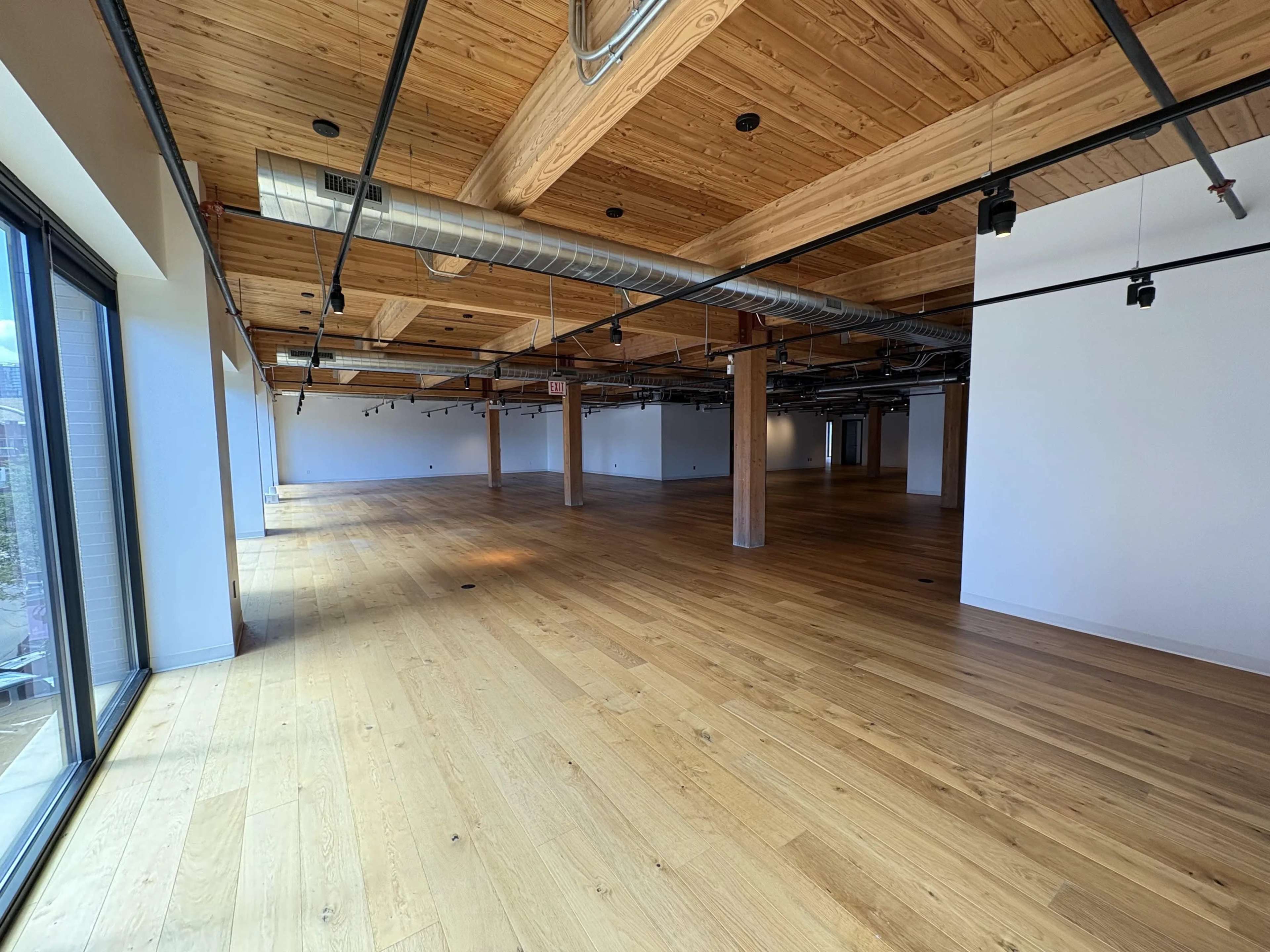 The image shows a spacious, empty room with wooden flooring, exposed beams, and large windows on one side.