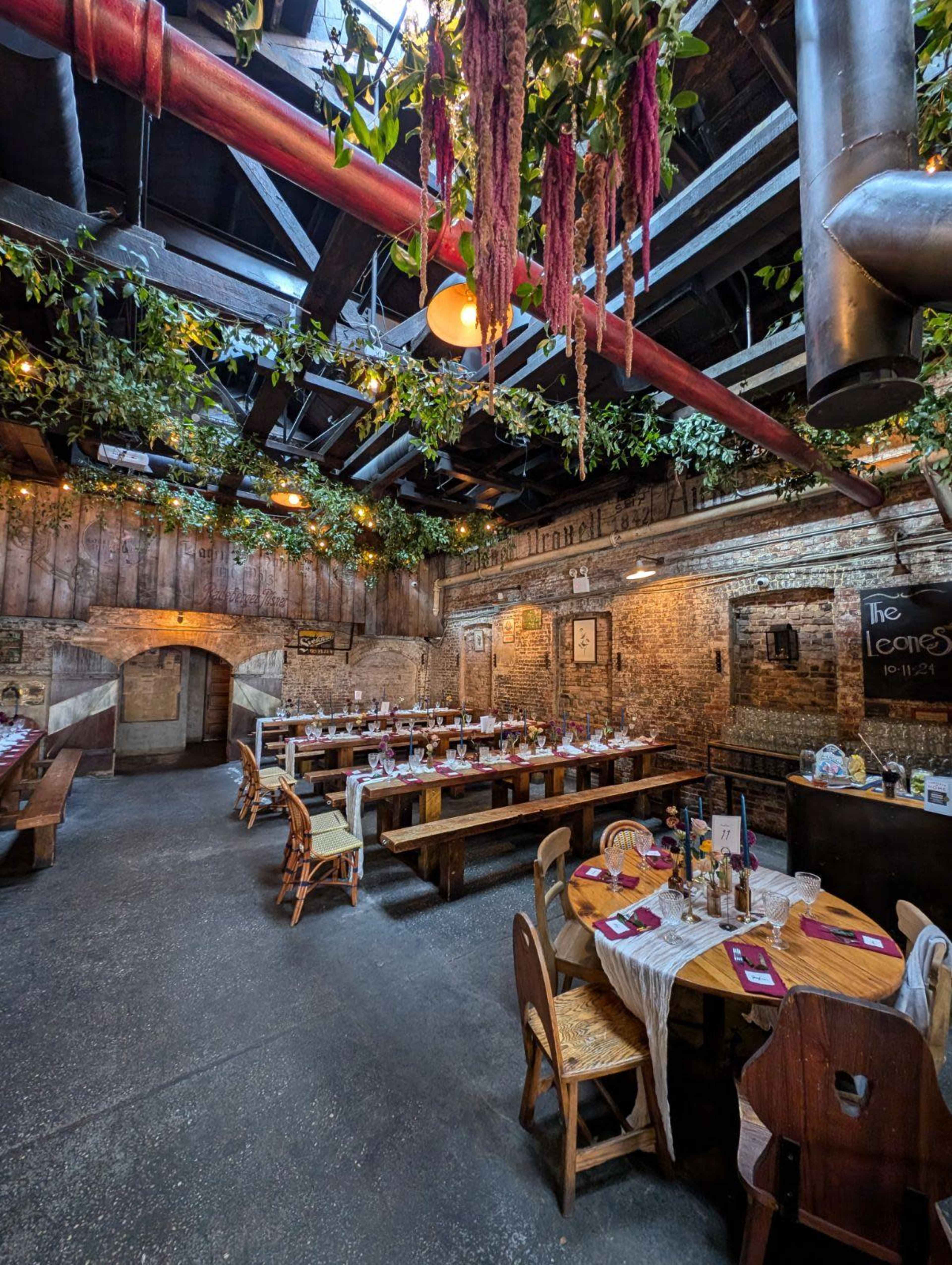 The image shows a rustic restaurant interior featuring long wooden tables set for dining, accented by hanging greenery and warm lighting.