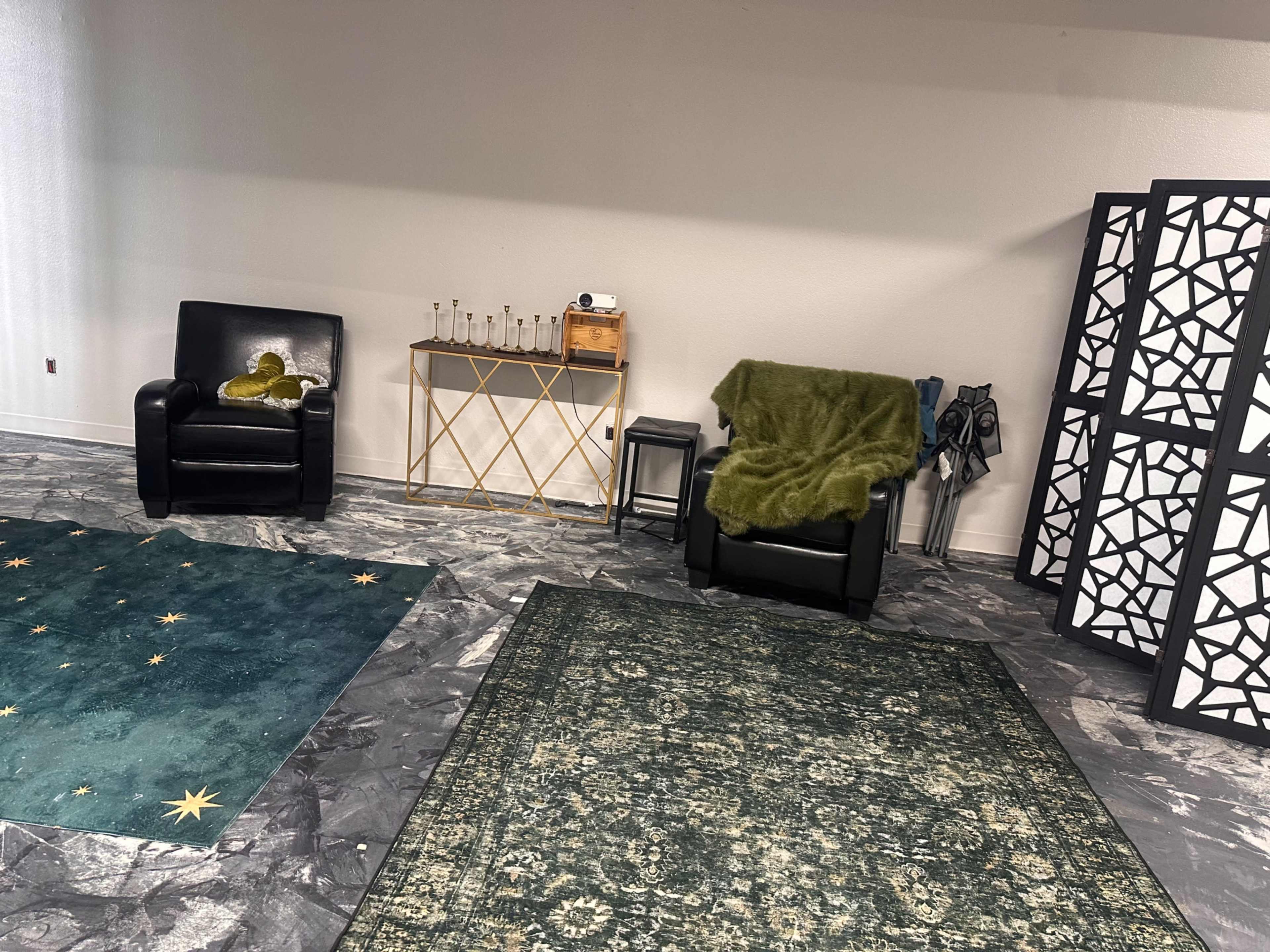 The image shows a room with two black chairs, a green blanket on one chair, a small table with glassware, and decorative dividers, all set against patterned rugs on a tiled floor.