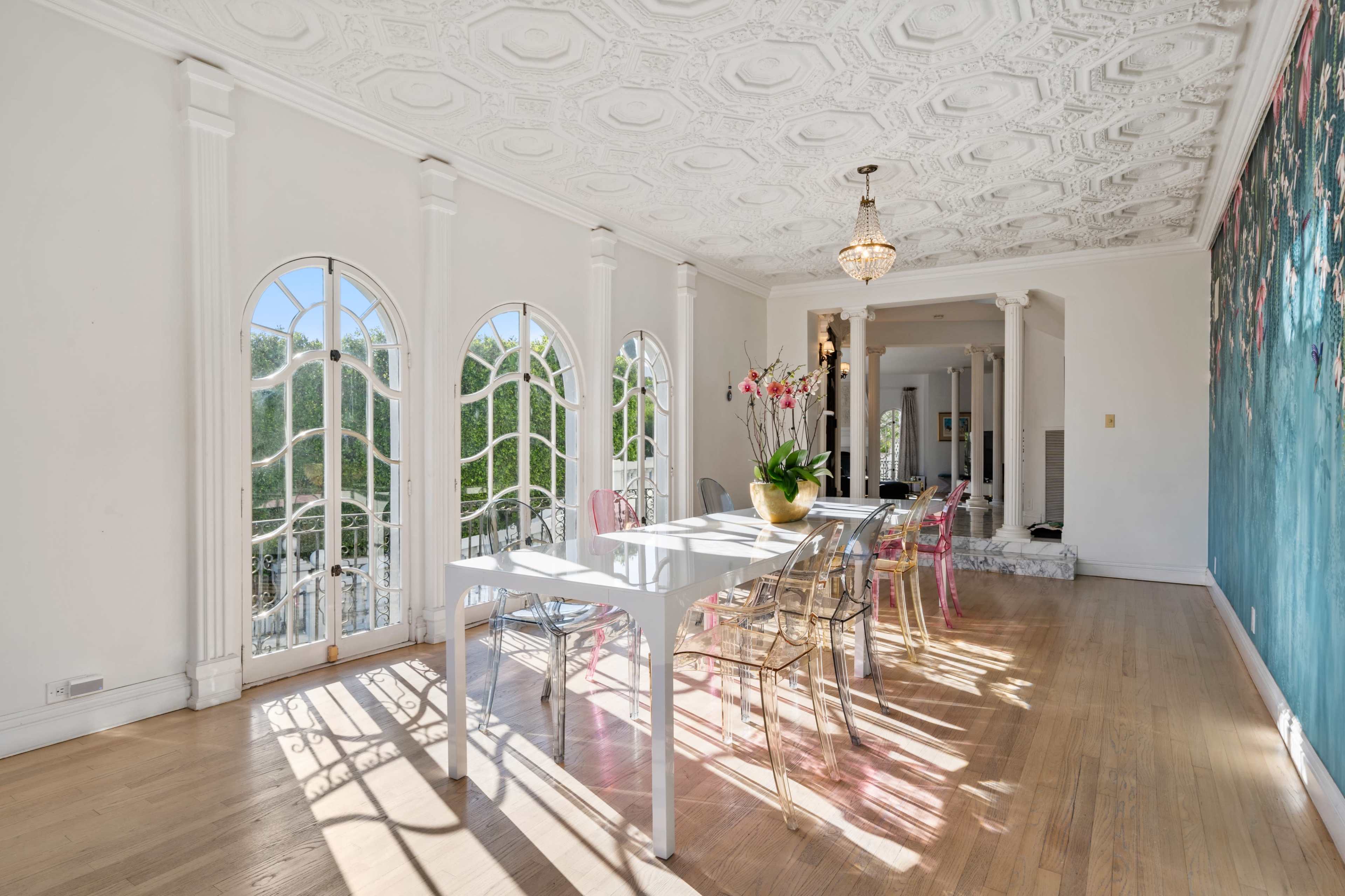 The image shows a bright dining area with a white table, clear plastic chairs, and large arched windows allowing natural light to fill the space.