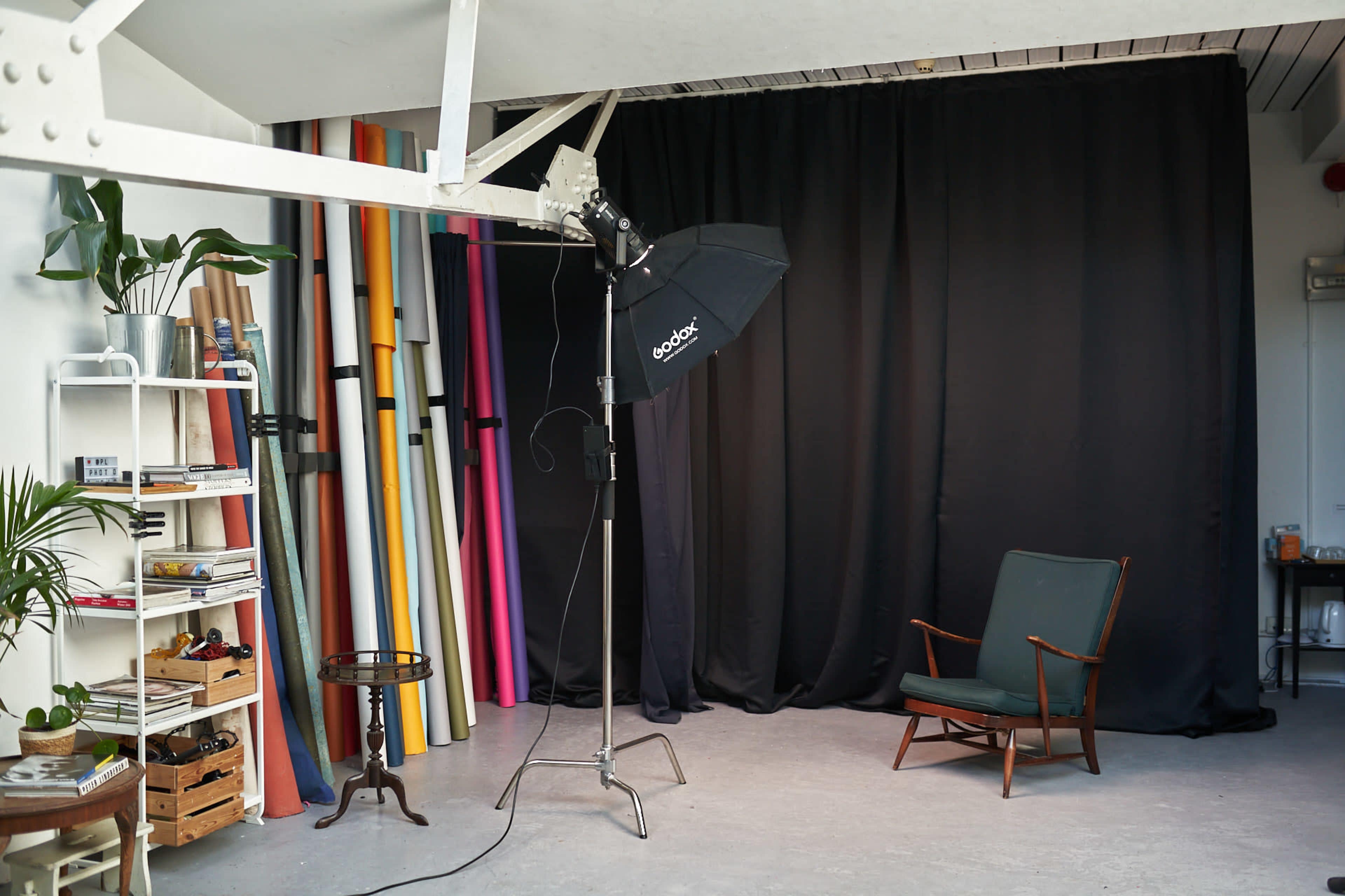 A photography studio with a black backdrop, a lighting setup, colorful rolls of paper, a chair, and shelves displaying various items.