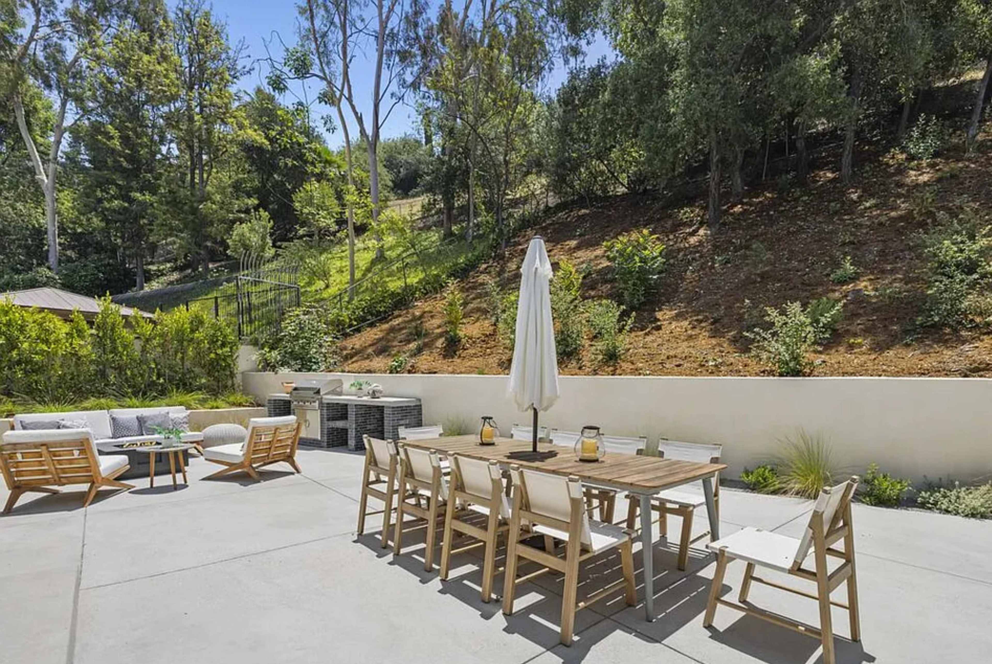 A patio features a large dining table surrounded by chairs, an umbrella, and a seating area with sofas, all set against a backdrop of trees and a hillside.