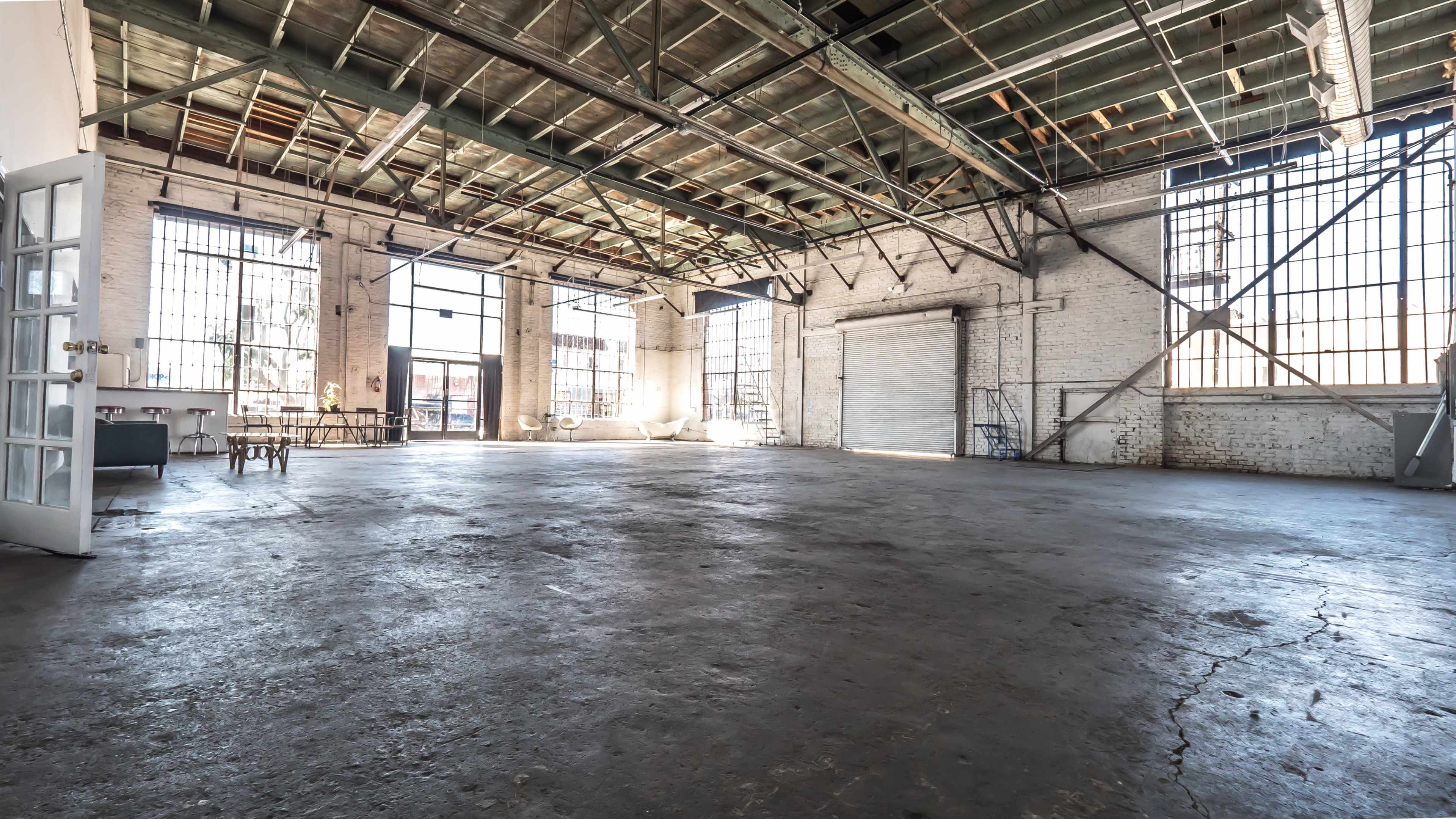 The image shows a large, empty industrial space with exposed beams, tall windows, and a concrete floor.