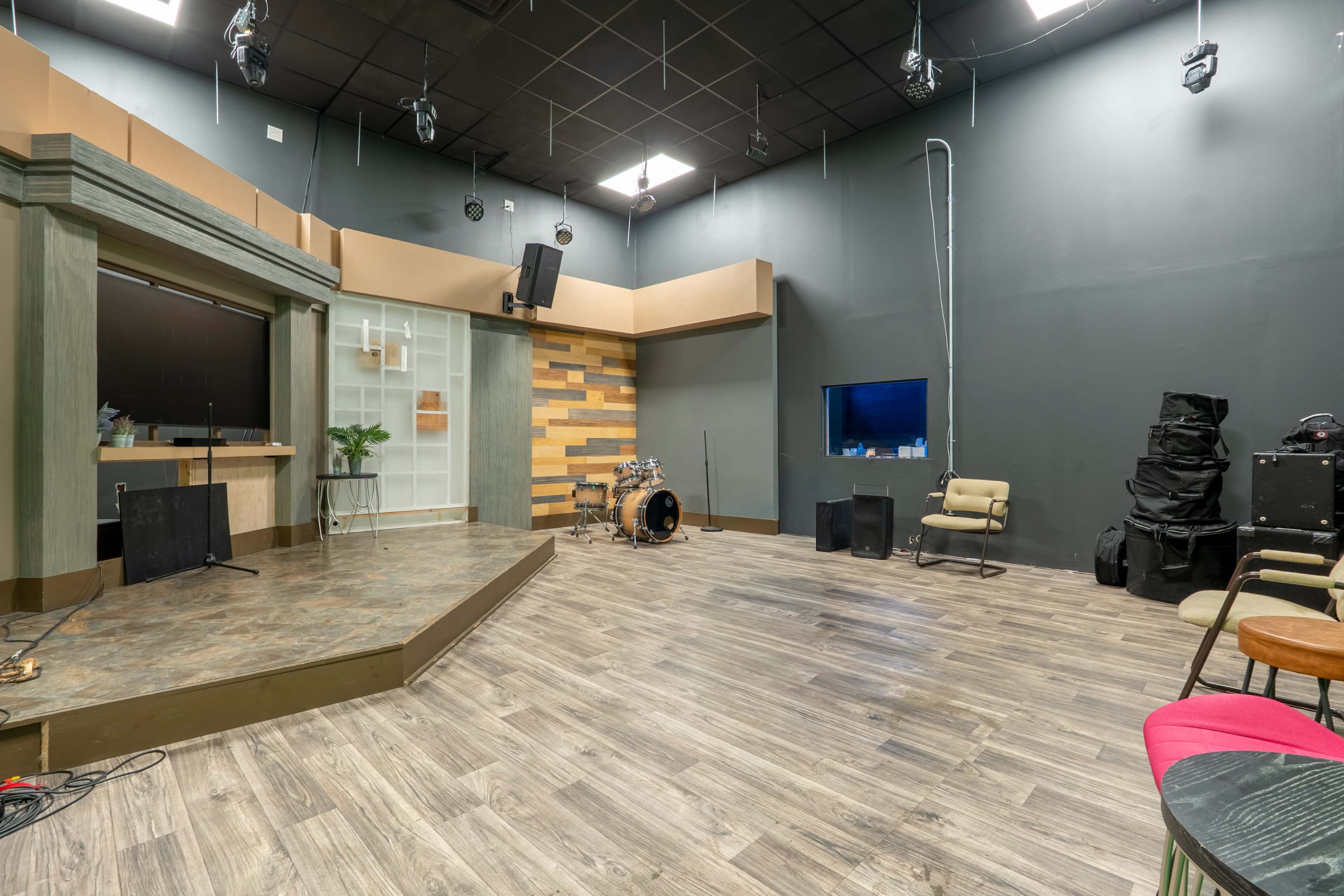 The image depicts a music studio with a wooden stage, seating area, and sound equipment, set against gray walls and a patterned backdrop.