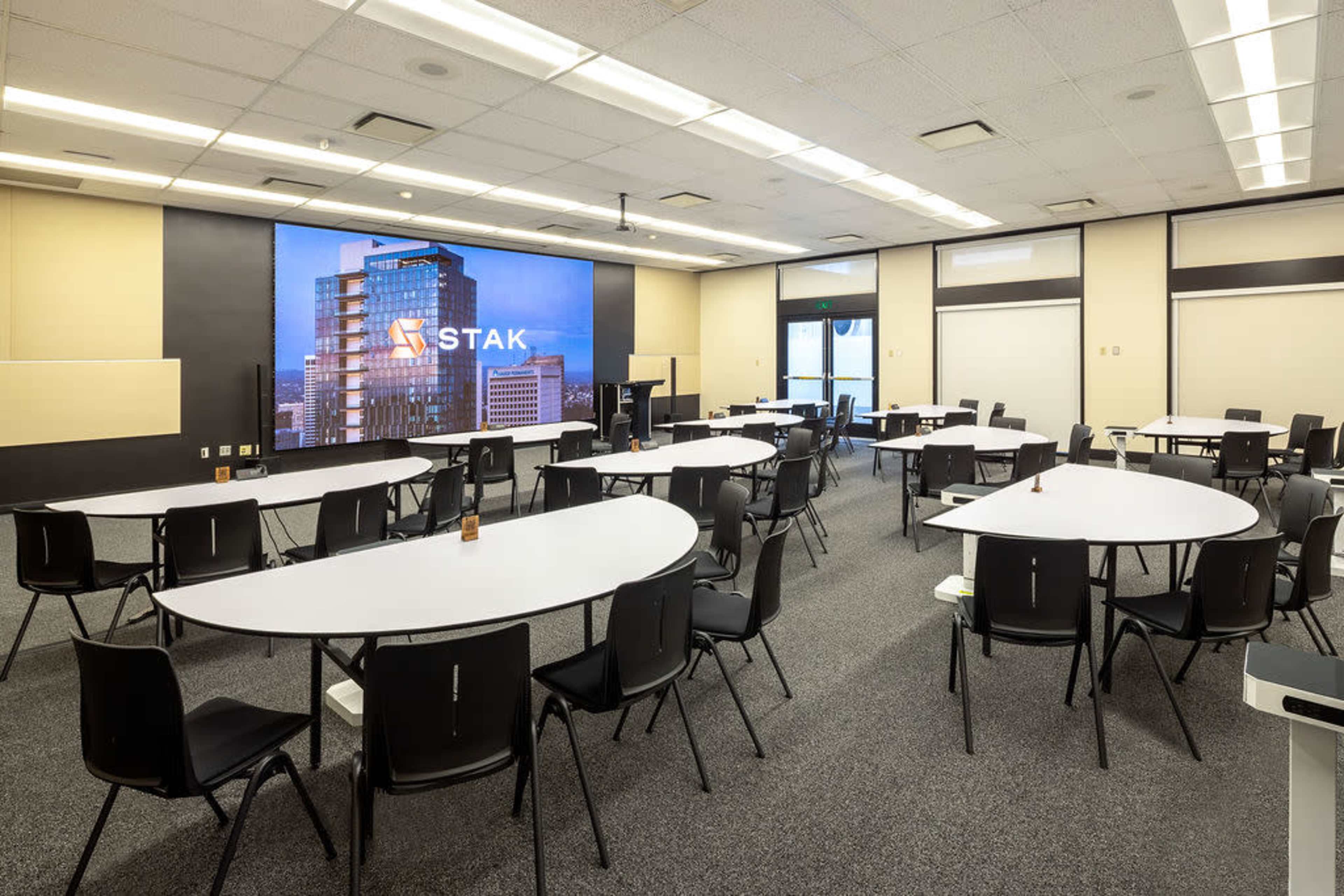 A conference room is arranged with several oval tables and black chairs, featuring a large screen displaying the logo "STAK" on one wall.