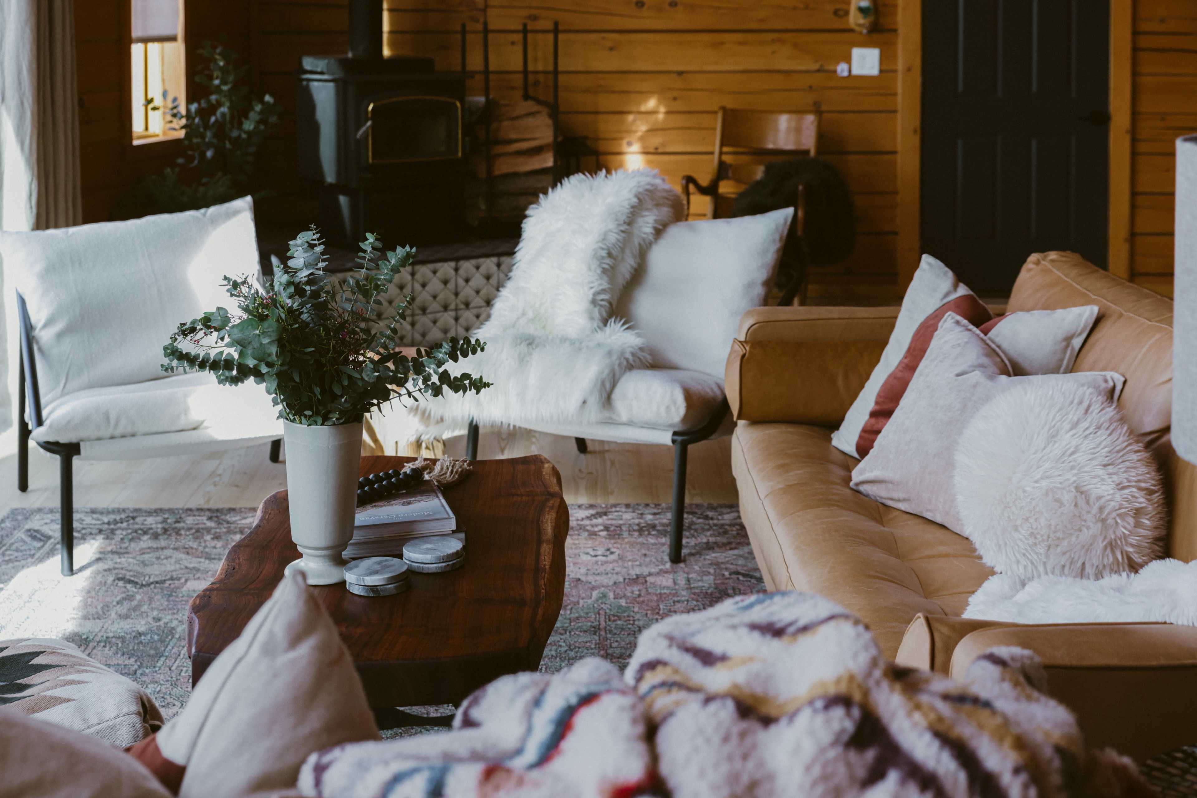 A cozy living room featuring a leather sofa, two white chairs, a wooden table, and a rustic wood stove, with a decorative plant and blankets scattered on the furniture.