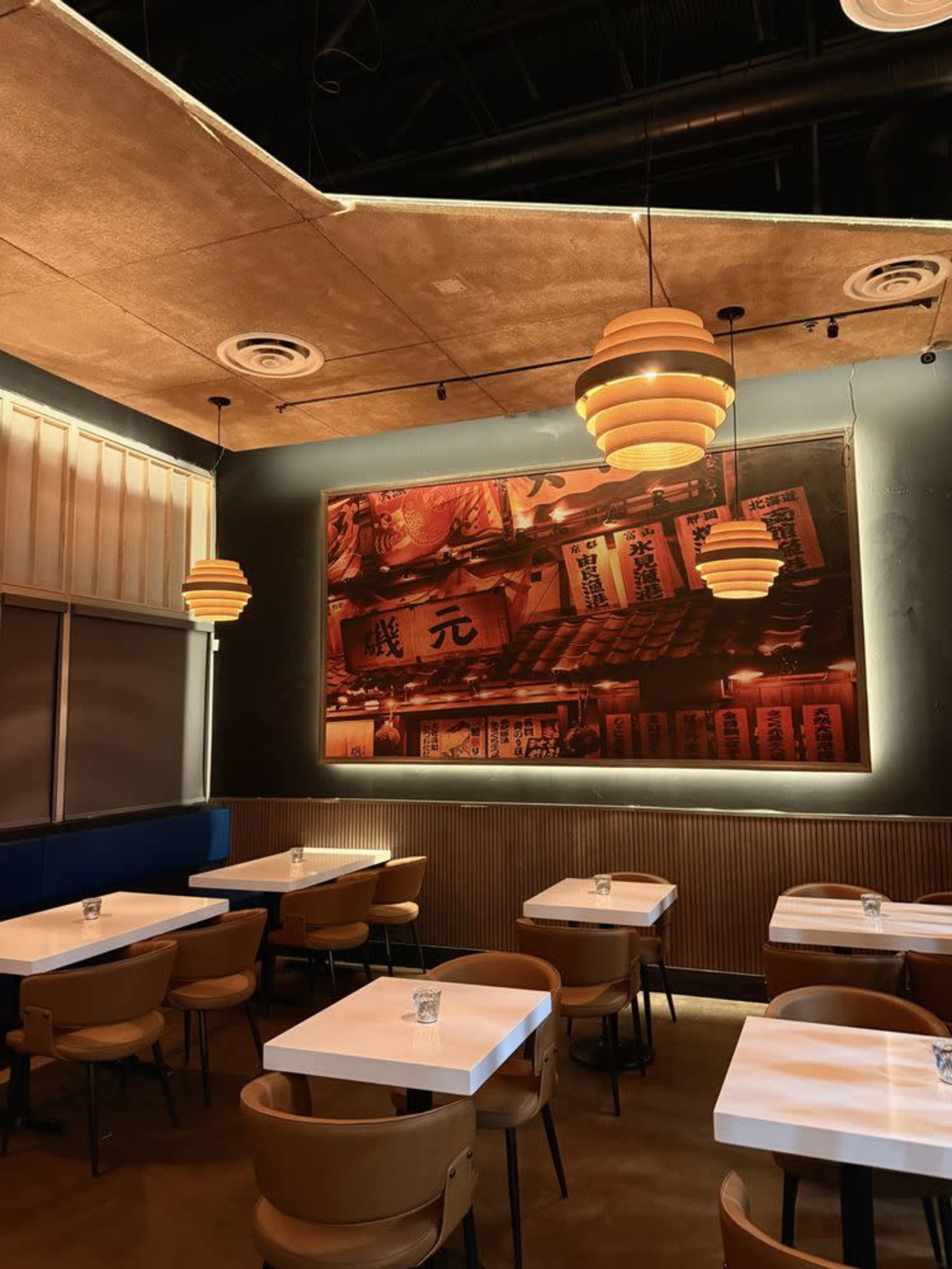 The image shows a restaurant interior with simple white tables, brown chairs, and an illuminated artwork on the wall.