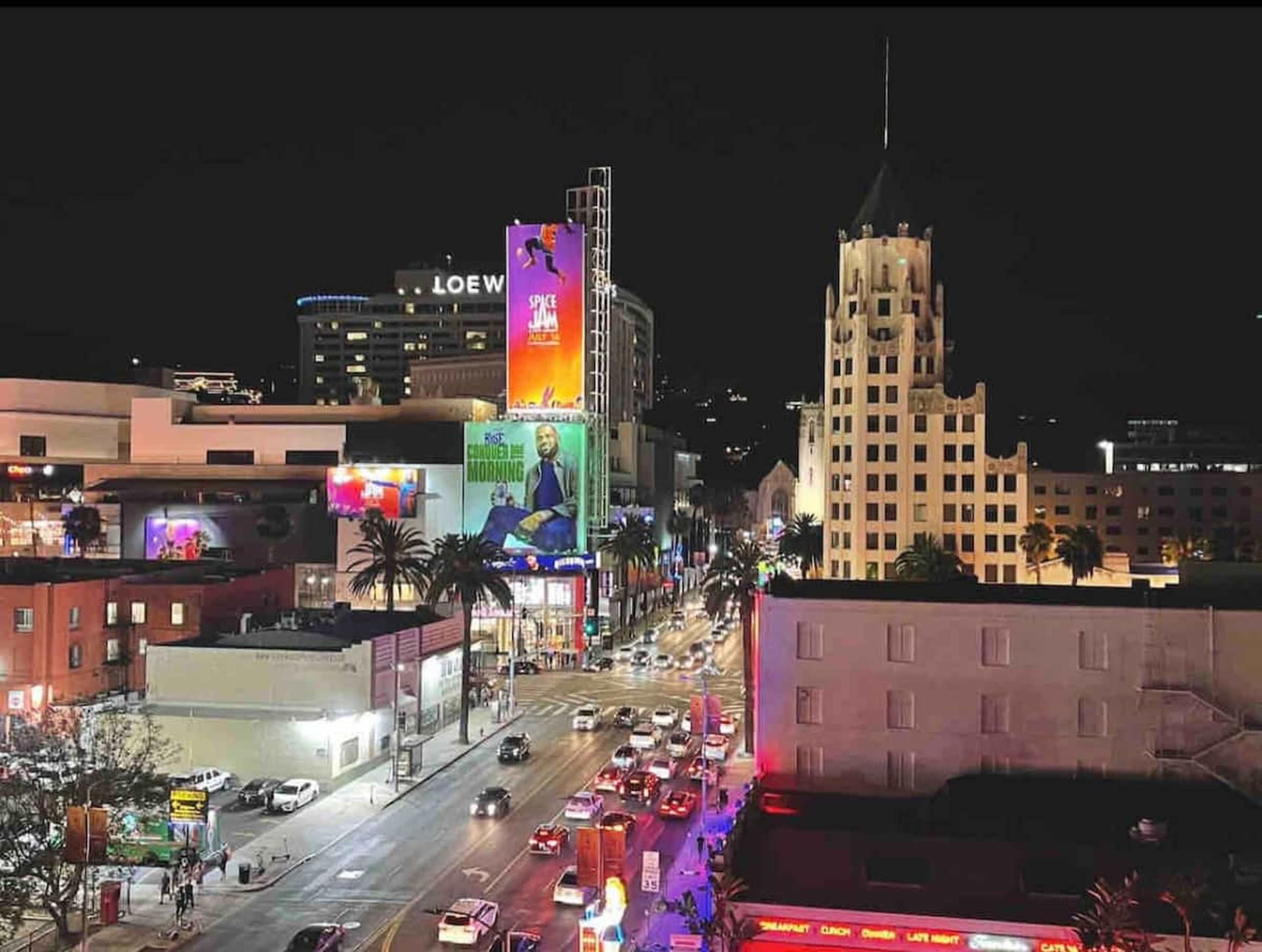 A vibrant cityscape of a busy Hollywood street at night, featuring illuminated billboards and palm trees.