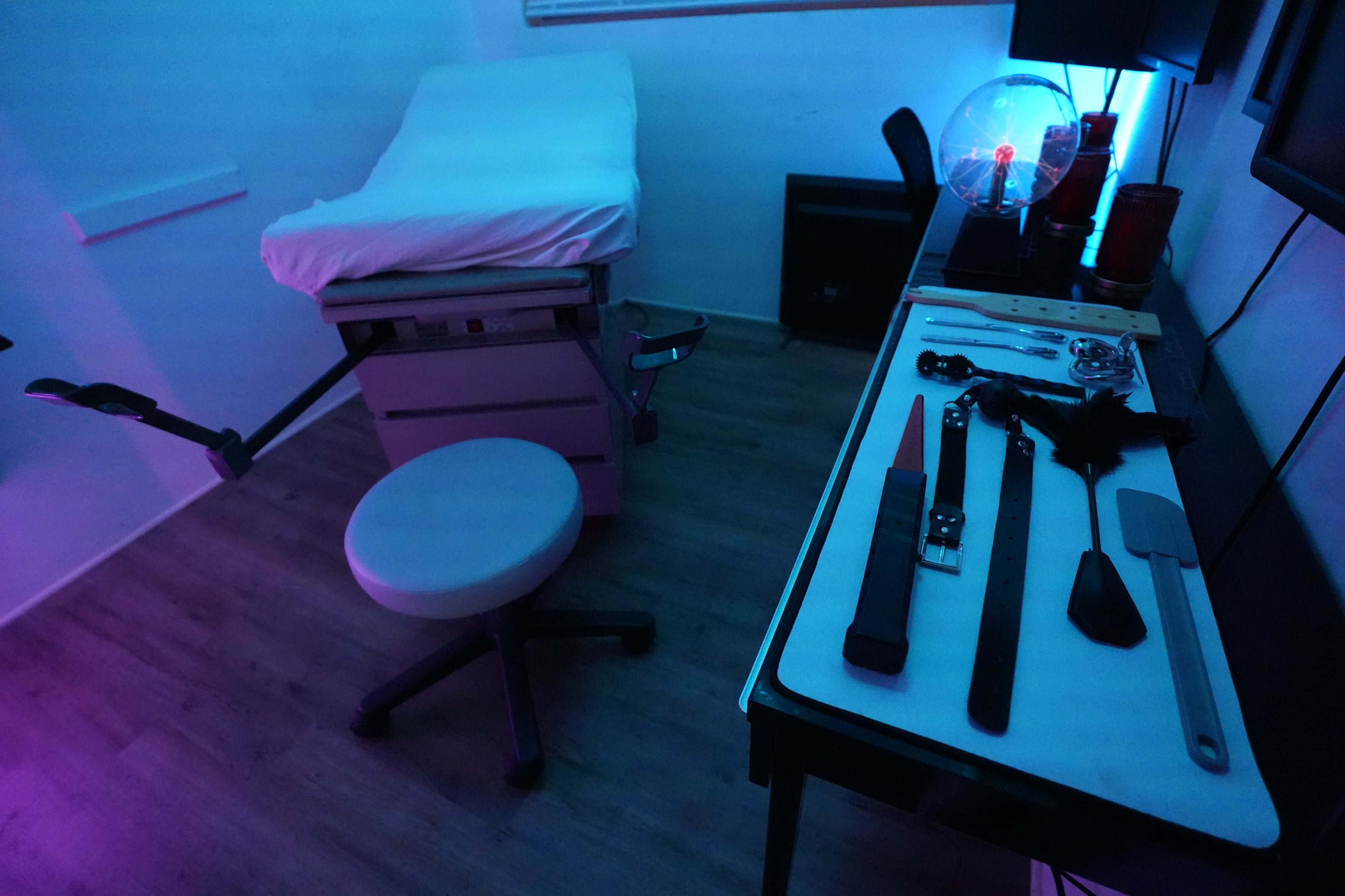 The image shows a dimly lit room with a medical examination table, a rolling stool, and a table displaying various tools and instruments.