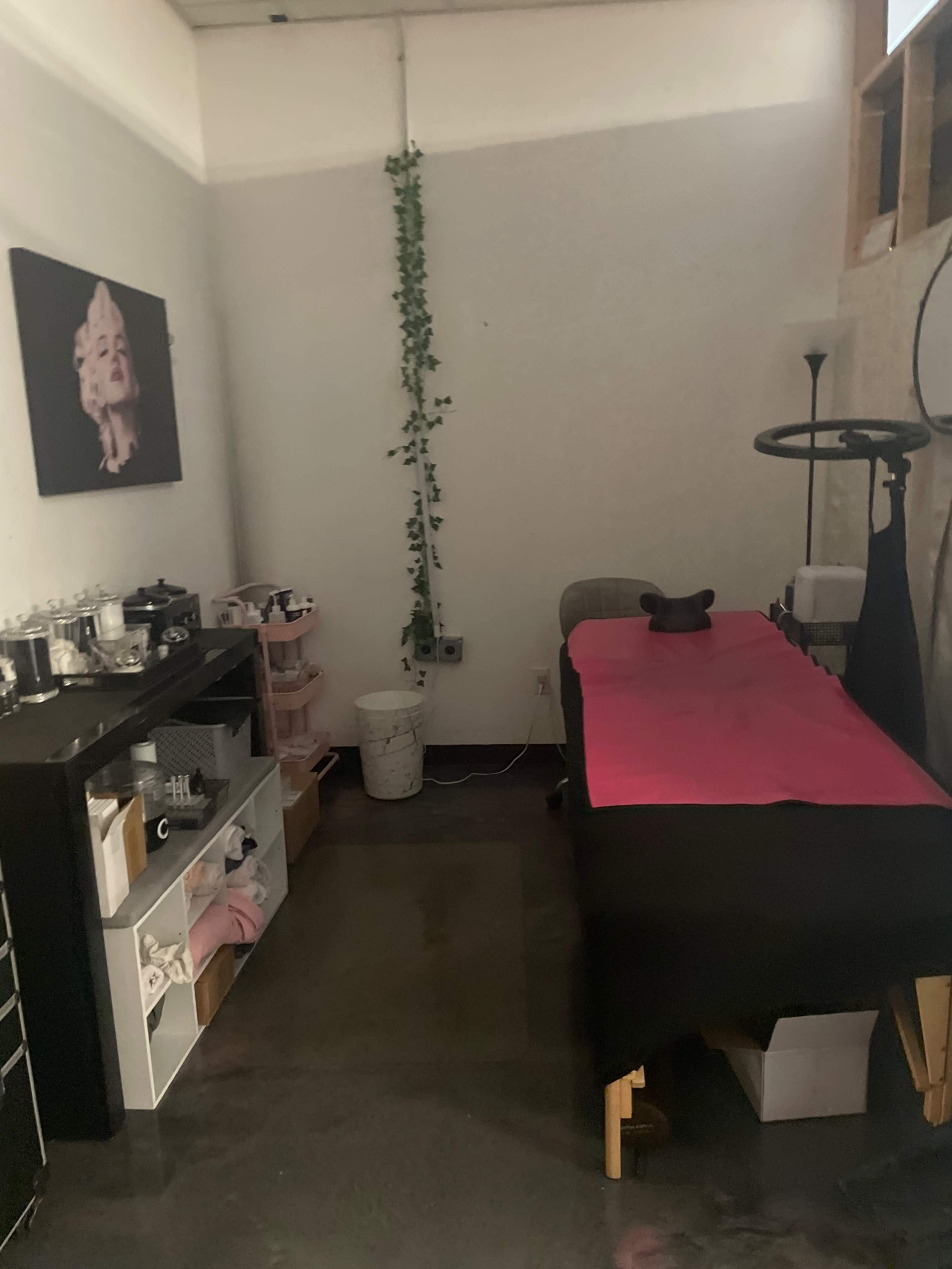 The image shows a small treatment room featuring a massage table covered with a pink sheet, a black cabinet with various items, and a wall adorned with a black-and-white portrait.
