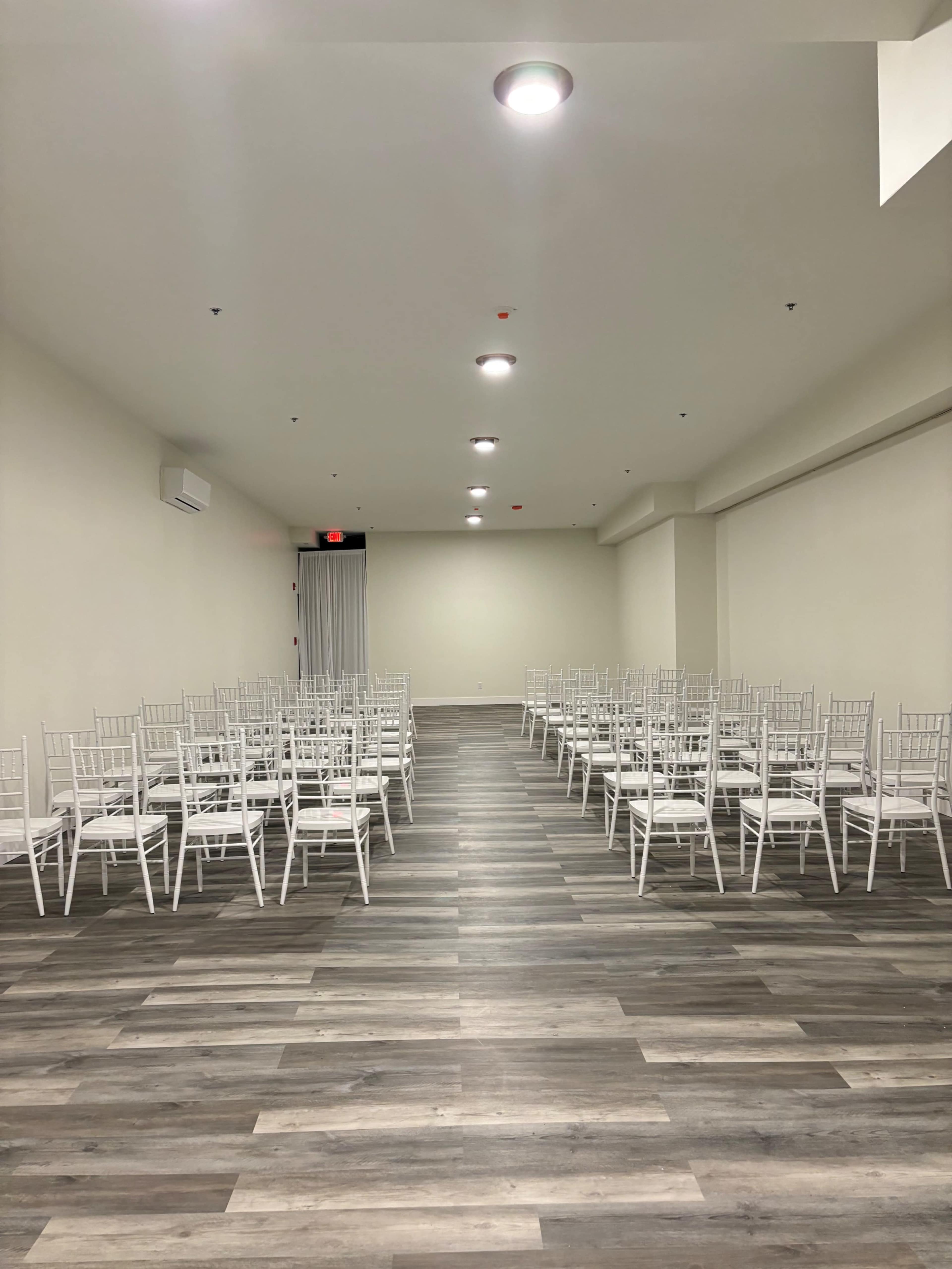 A spacious room with rows of white chiavari chairs arranged on a wooden floor.