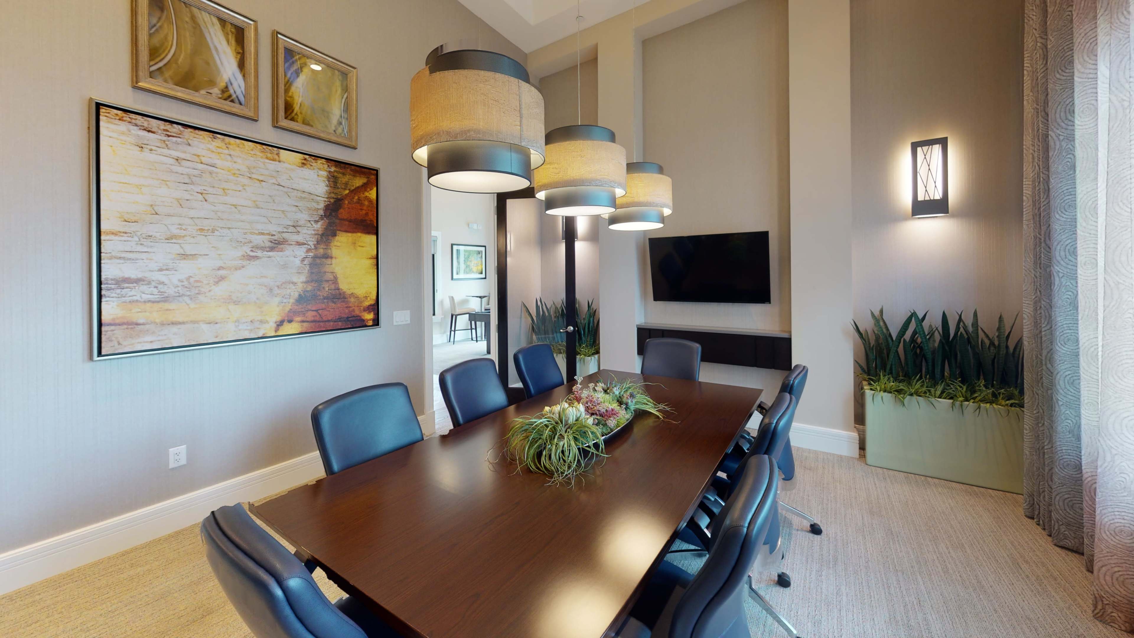 A modern conference room with a large wooden table surrounded by black chairs, featuring pendant lights, artwork on the walls, and a television.
