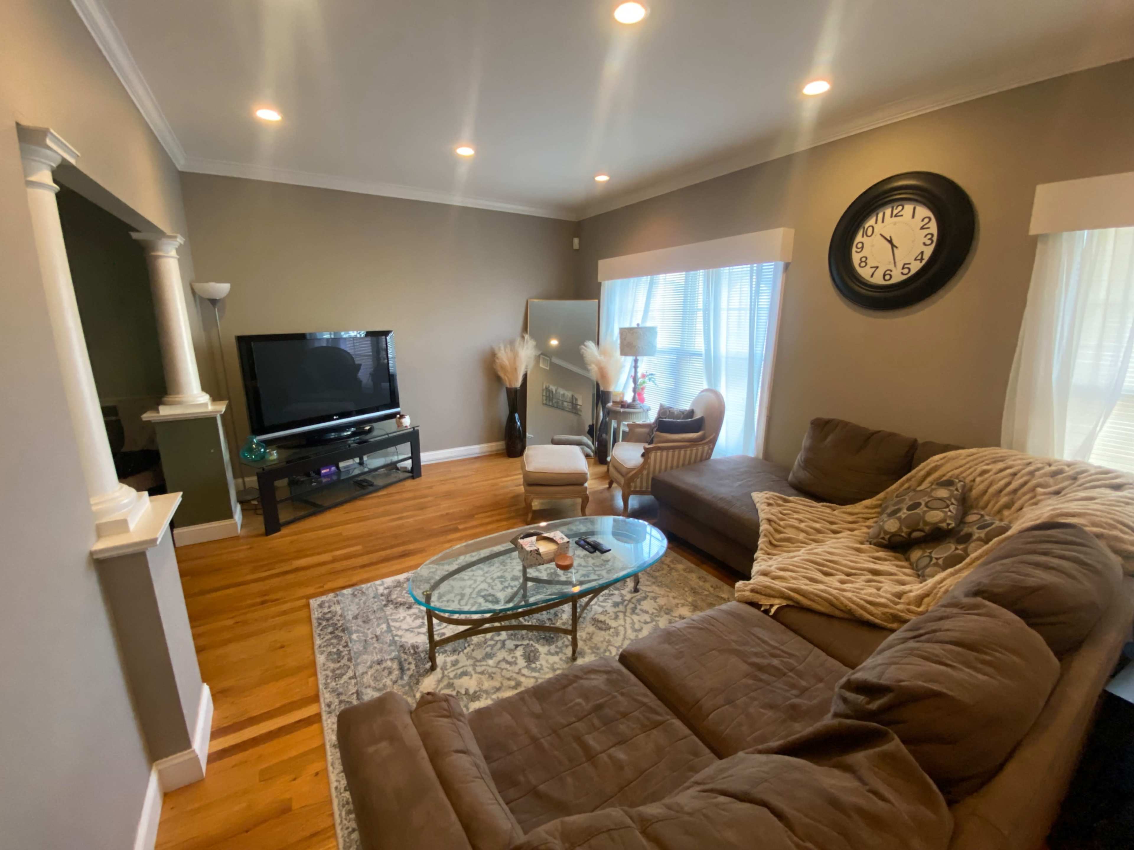 The image shows a cozy living room with a sectional sofa, a coffee table, a television, and large windows allowing natural light to enter.