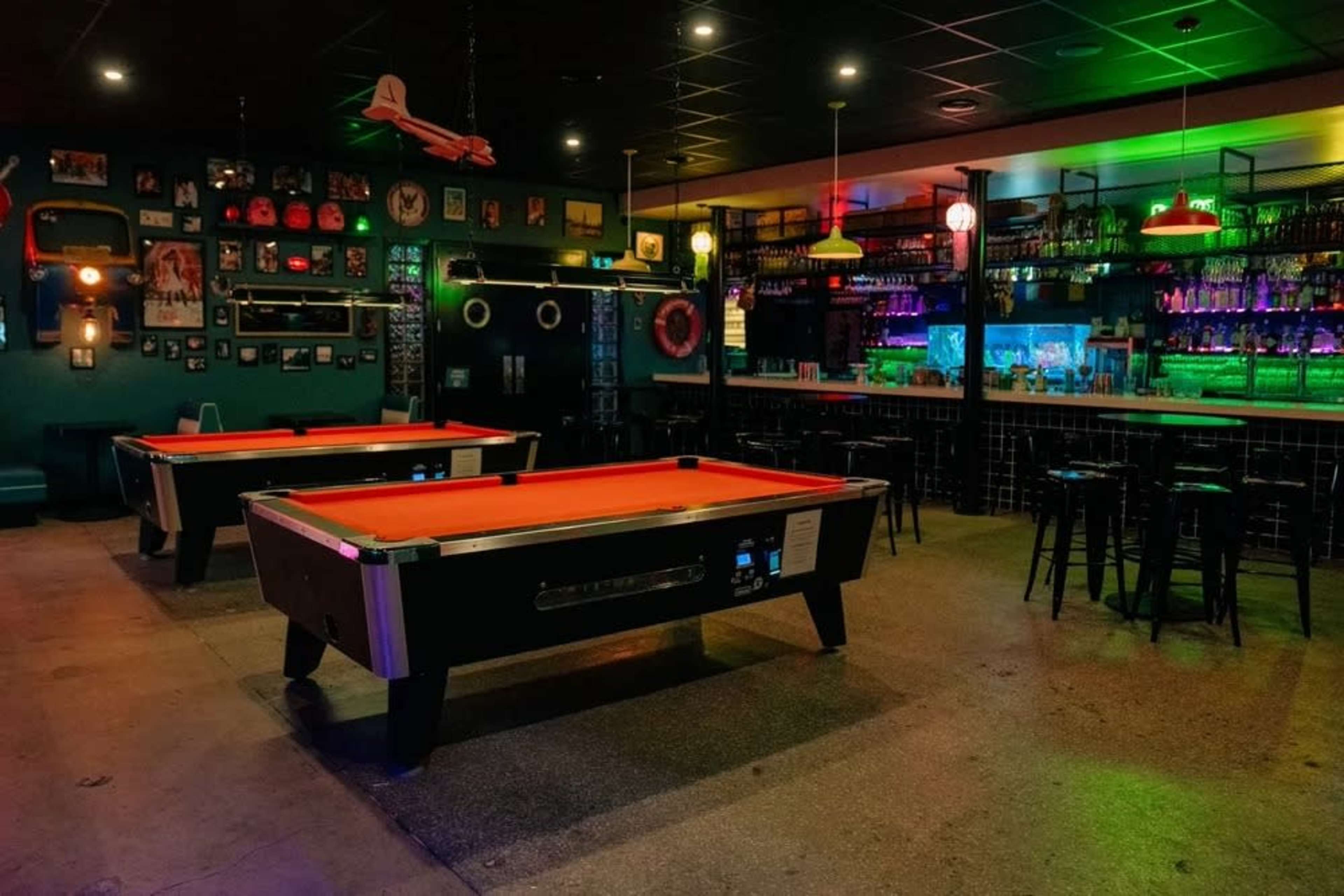 The image shows a dimly lit bar with two billiard tables and a colorful backlit bar area.