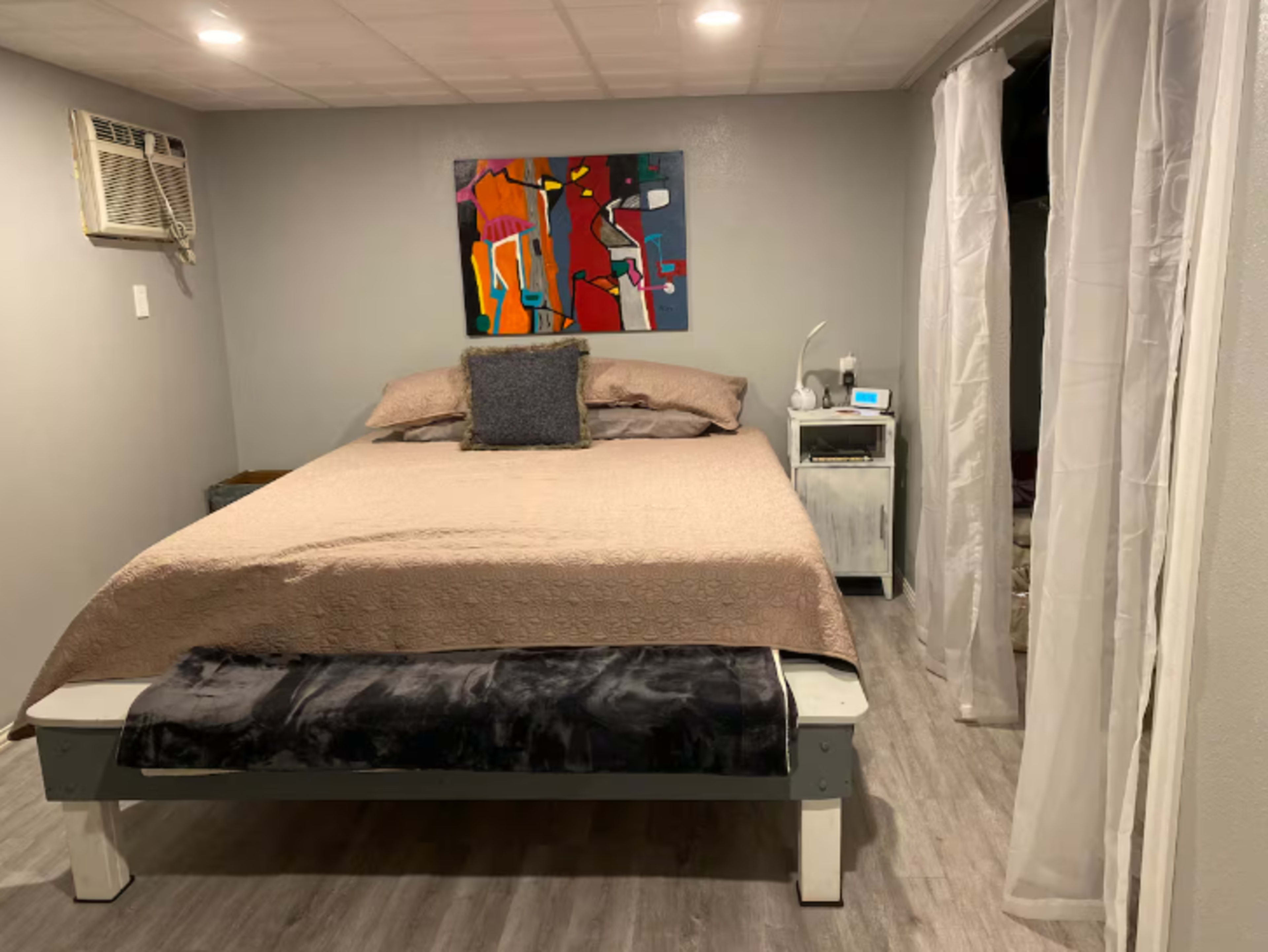 The image shows a bedroom with a double bed covered in a beige blanket, a colorful painting on the wall, and an air conditioning unit above the bed.