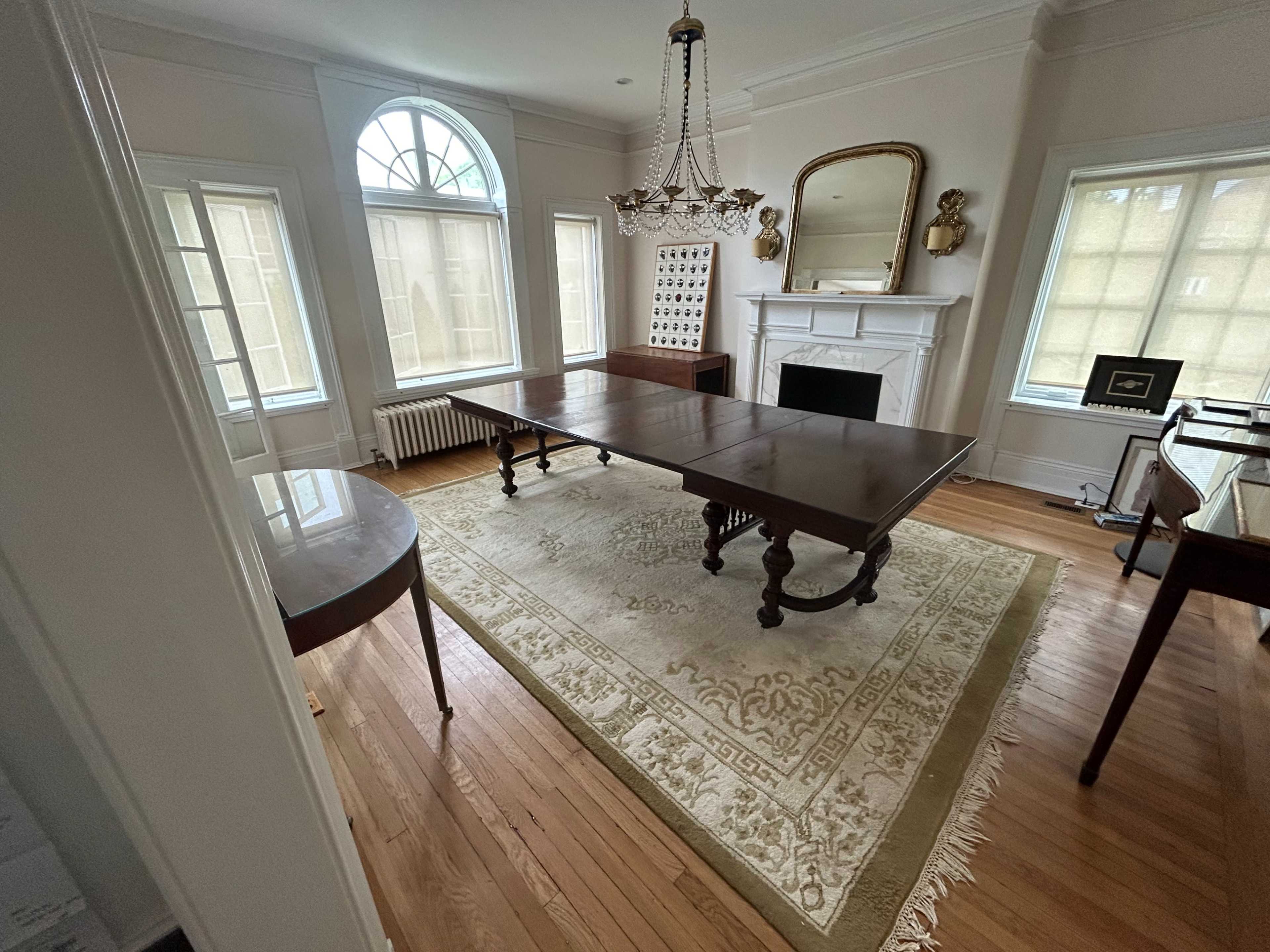 The image shows a spacious room with a large wooden table, a chandelier, and a fireplace, surrounded by windows.