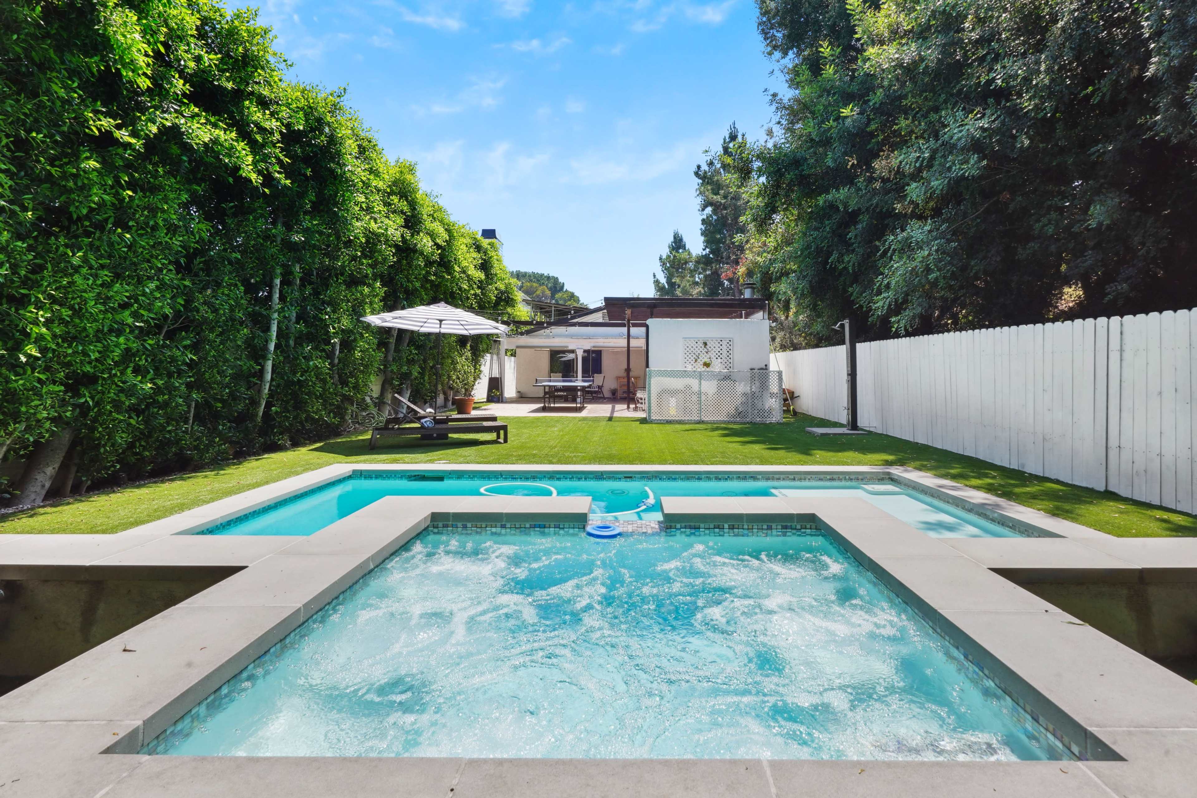 A clear swimming pool and hot tub are set in a backyard surrounded by lush greenery and a white fence, with a house and patio visible in the background.