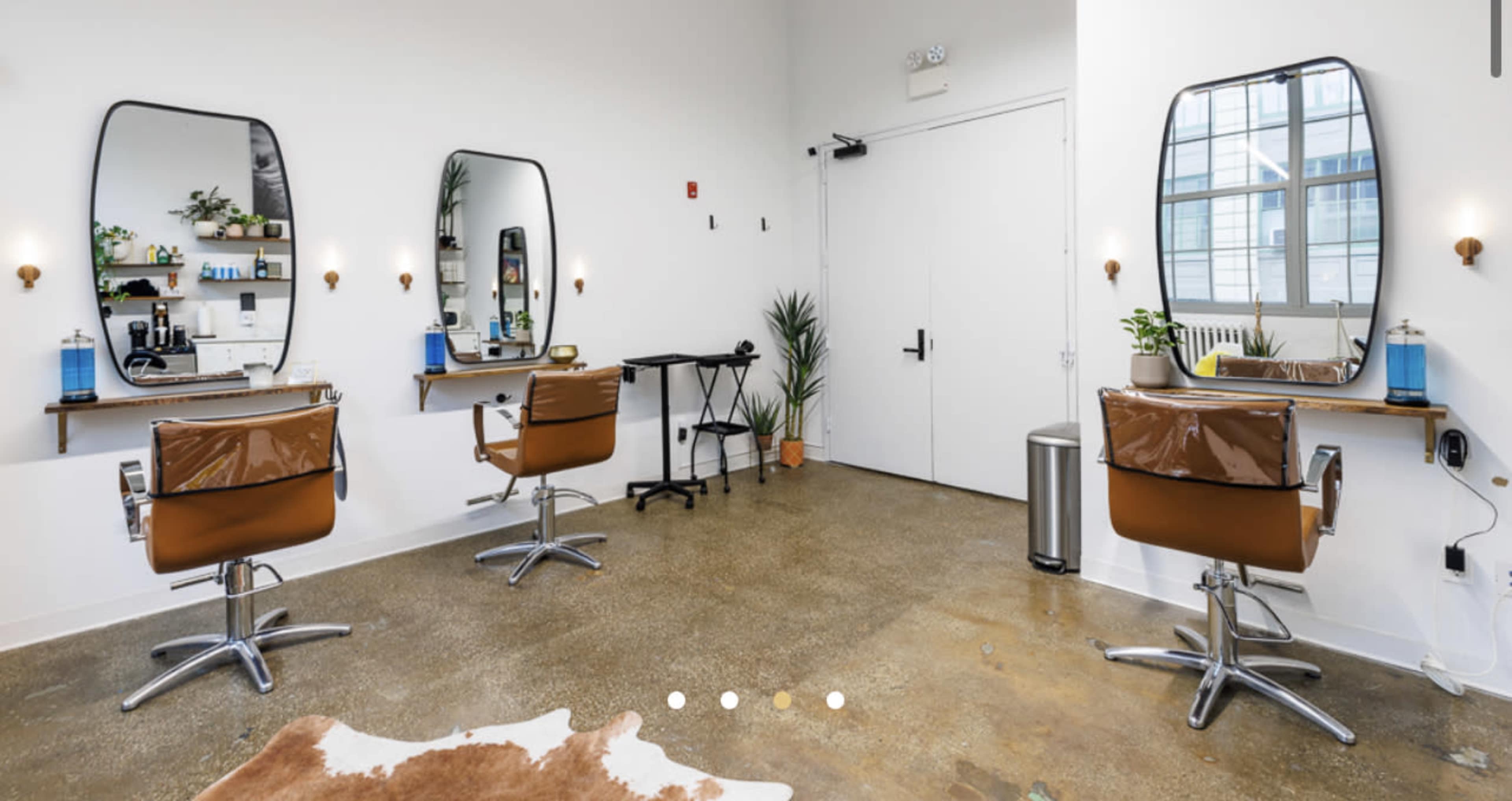 A hair salon with three styling stations, each featuring a brown chair and a large mirror, set against a minimalist interior.