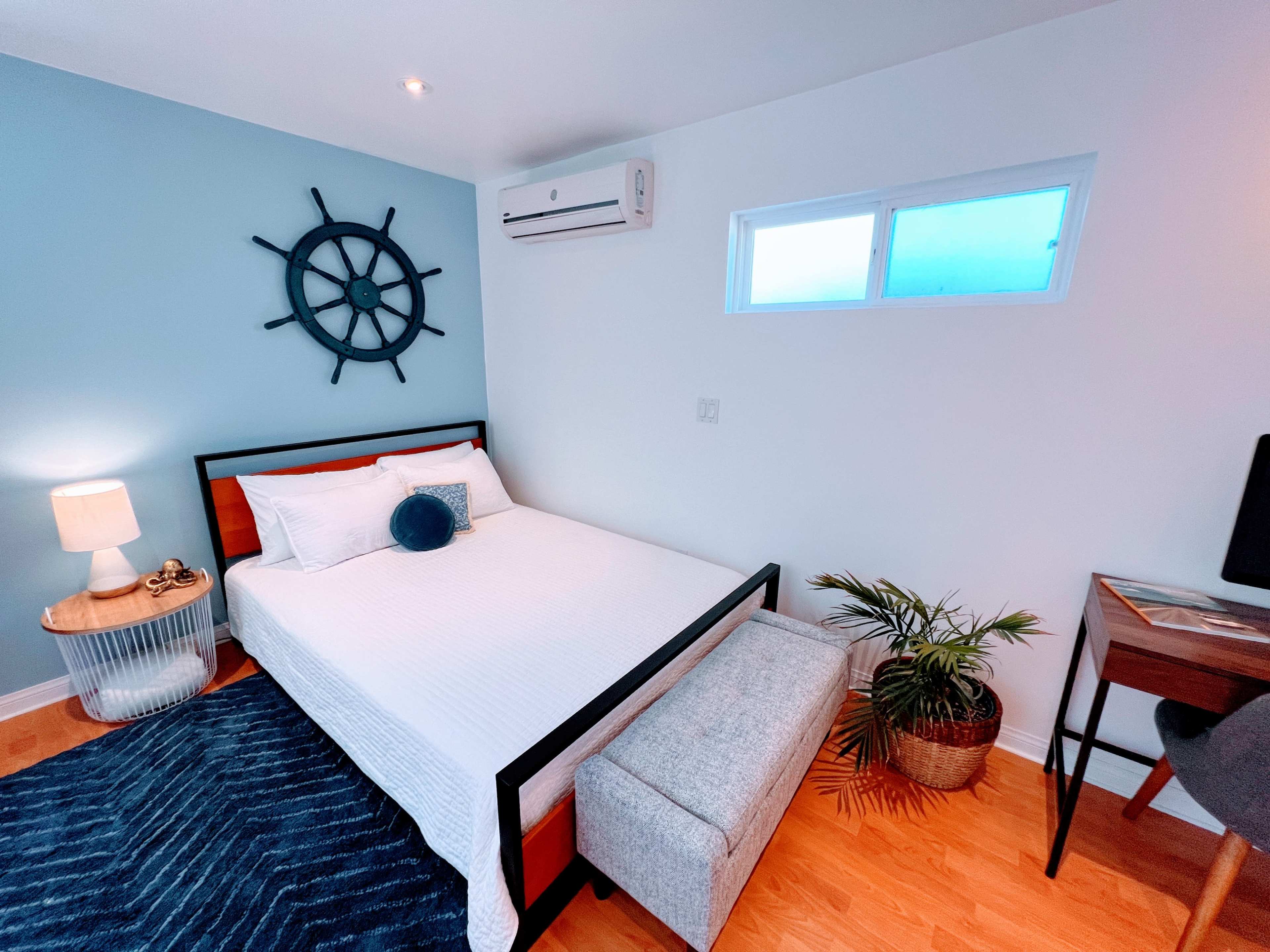 A neatly arranged bedroom featuring a bed, a small side table with a lamp, a decorative wall piece, a potted plant, and a desk in the corner.